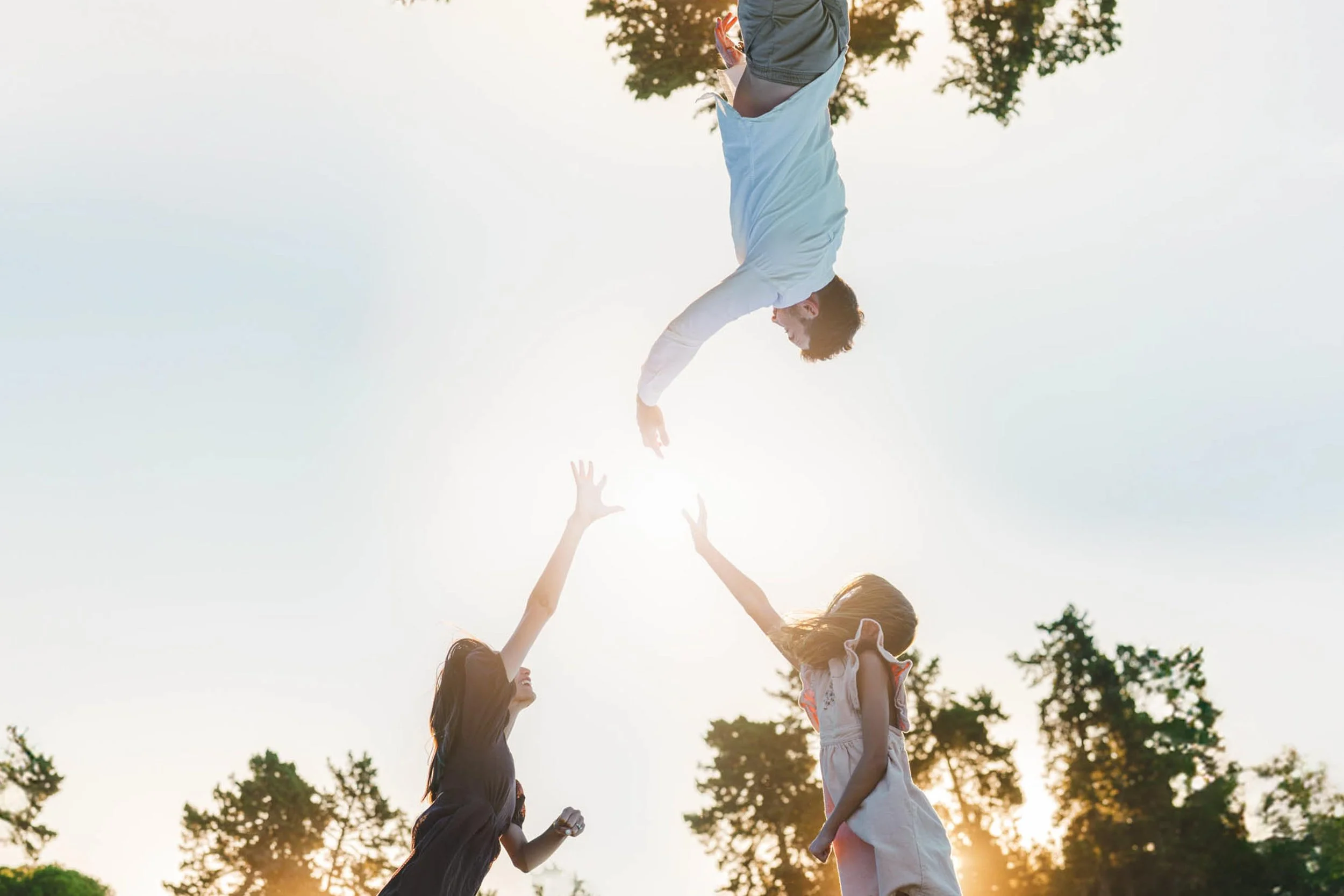 A man appears to be falling from the sky towards two women with trees and a bright sky in the background.