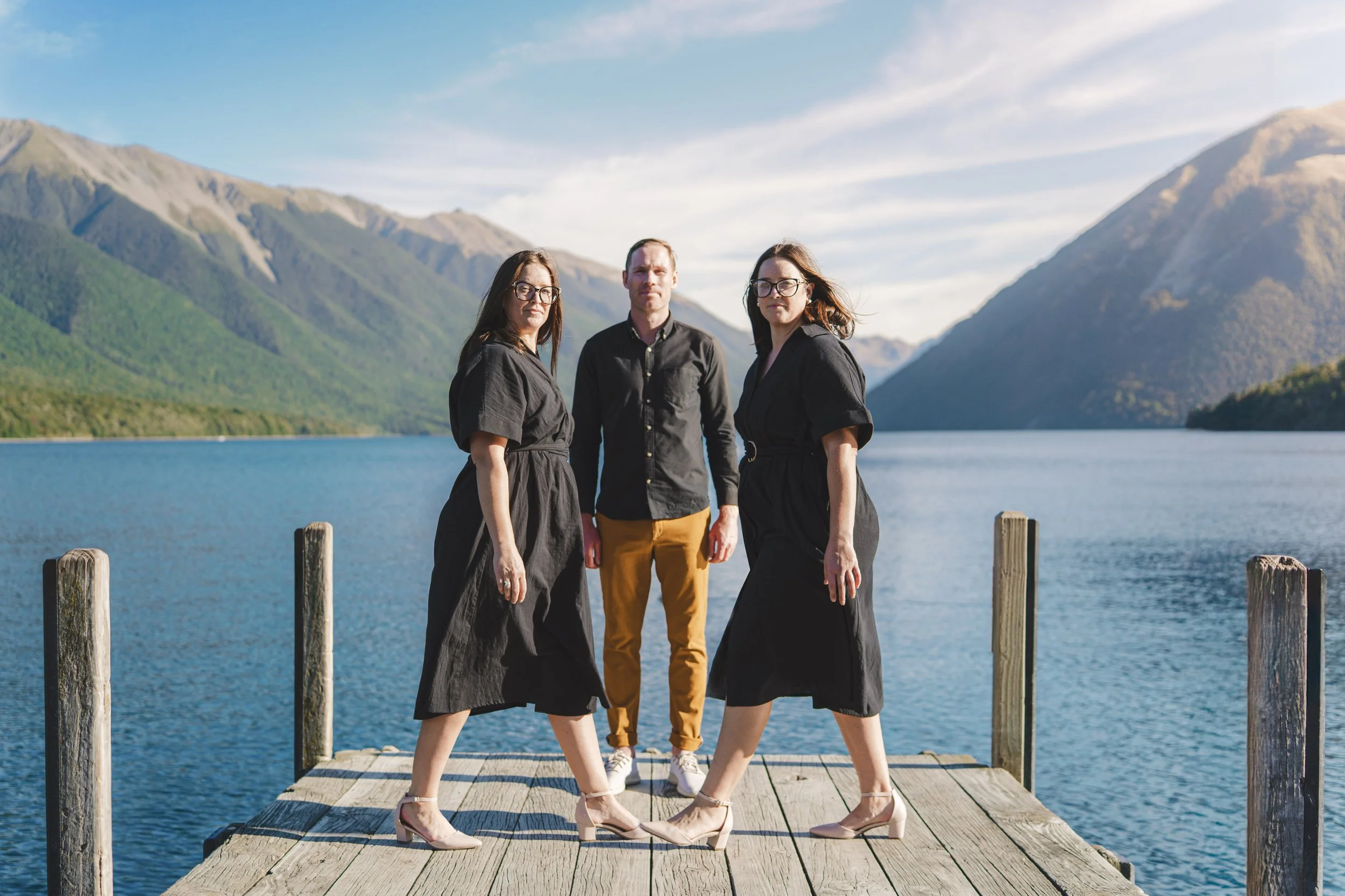 Three people standing on a wooden dock by a lake with mountains in the background. The two women are wearing black dresses and heels, and the man is wearing a black shirt and tan pants.