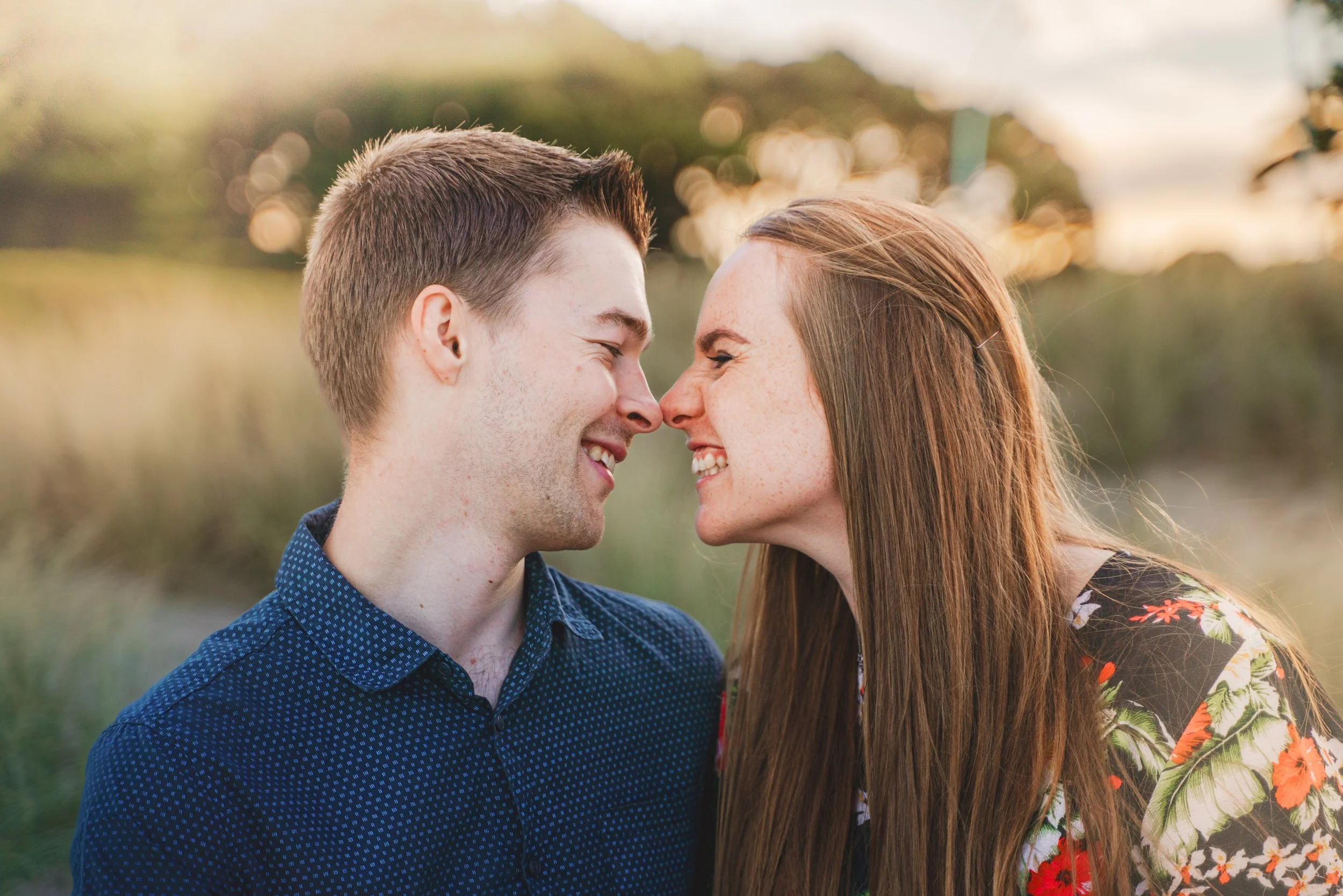 A young man and woman nose-to-nose, smiling happily outdoors during sunset, with blurred natural background.