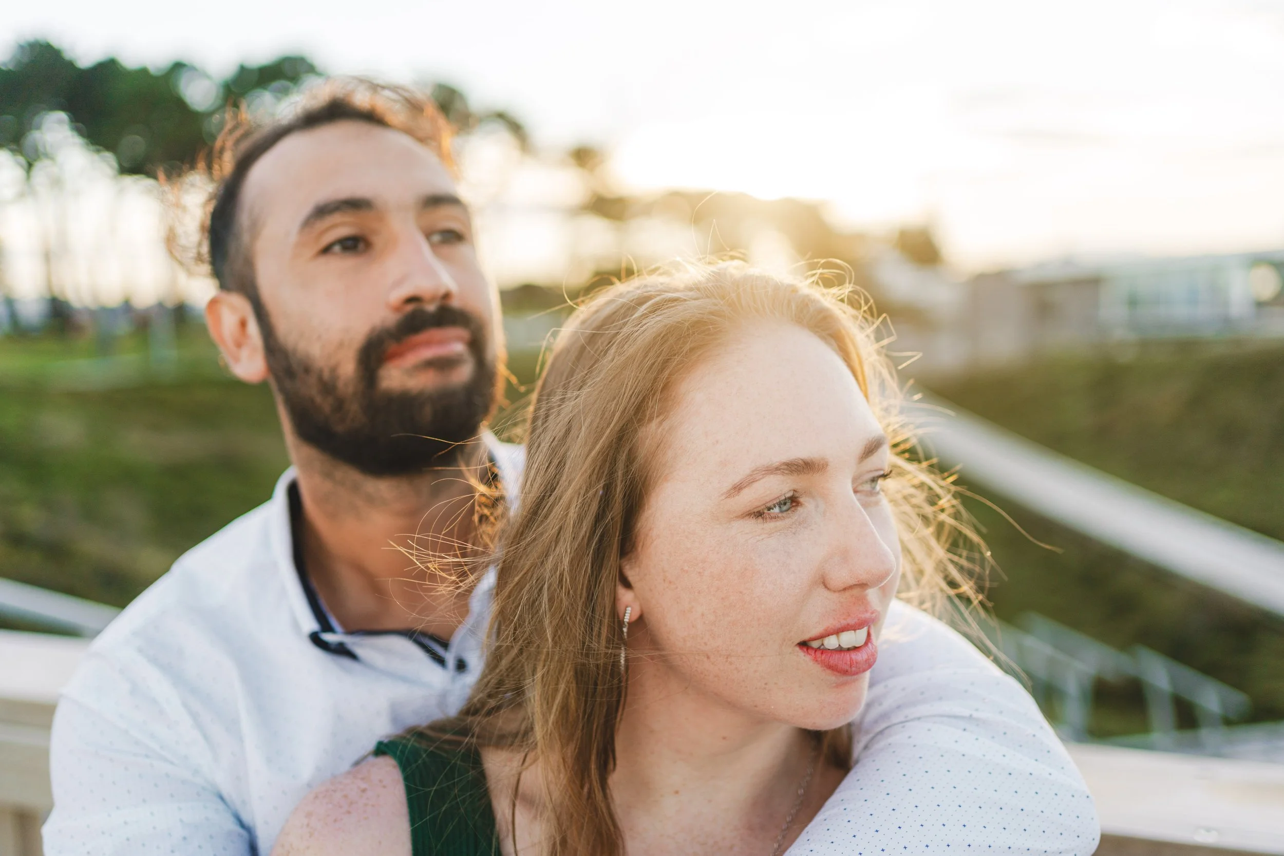 A couple sitting outdoors during sunset, with the man behind the woman, both looking into the distance.
