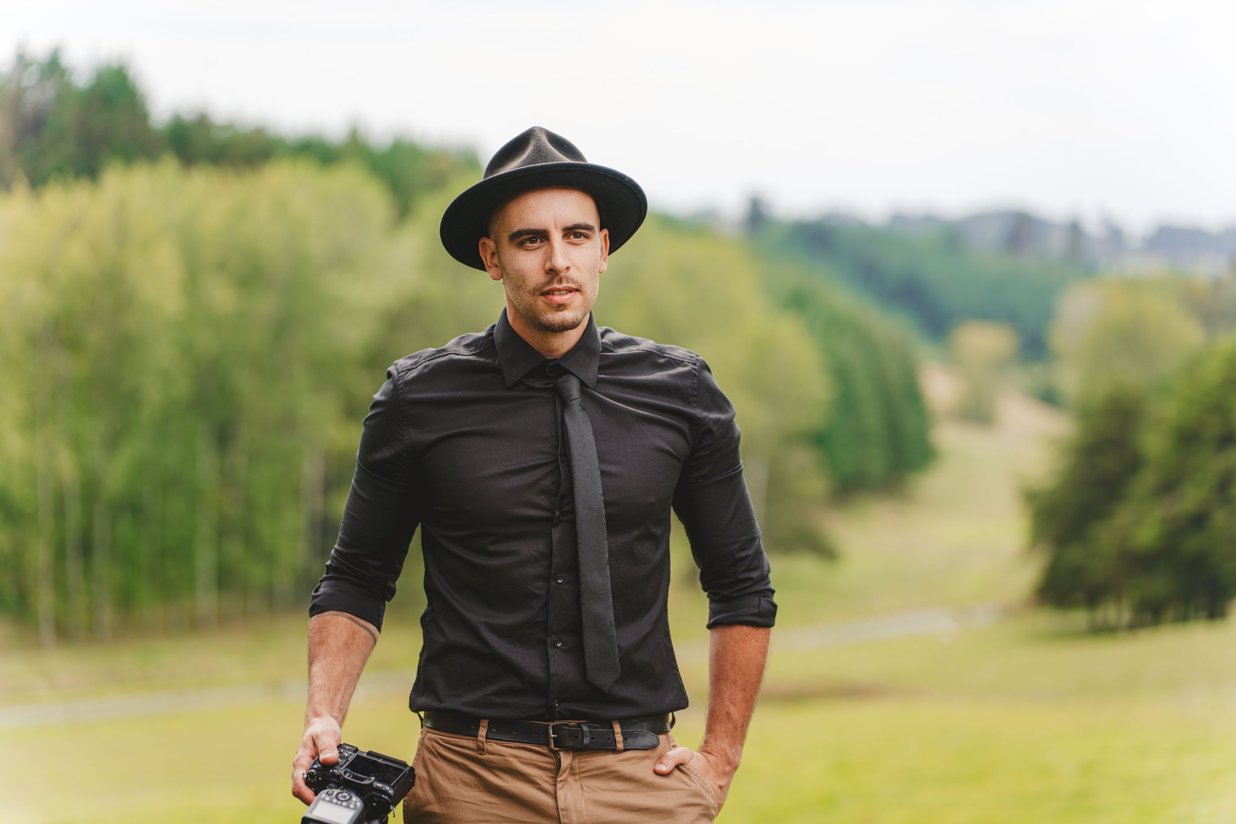 A man wearing a black shirt, black tie, and black hat standing outdoors in a green landscape, holding a camera in his right hand, with a thoughtful expression.