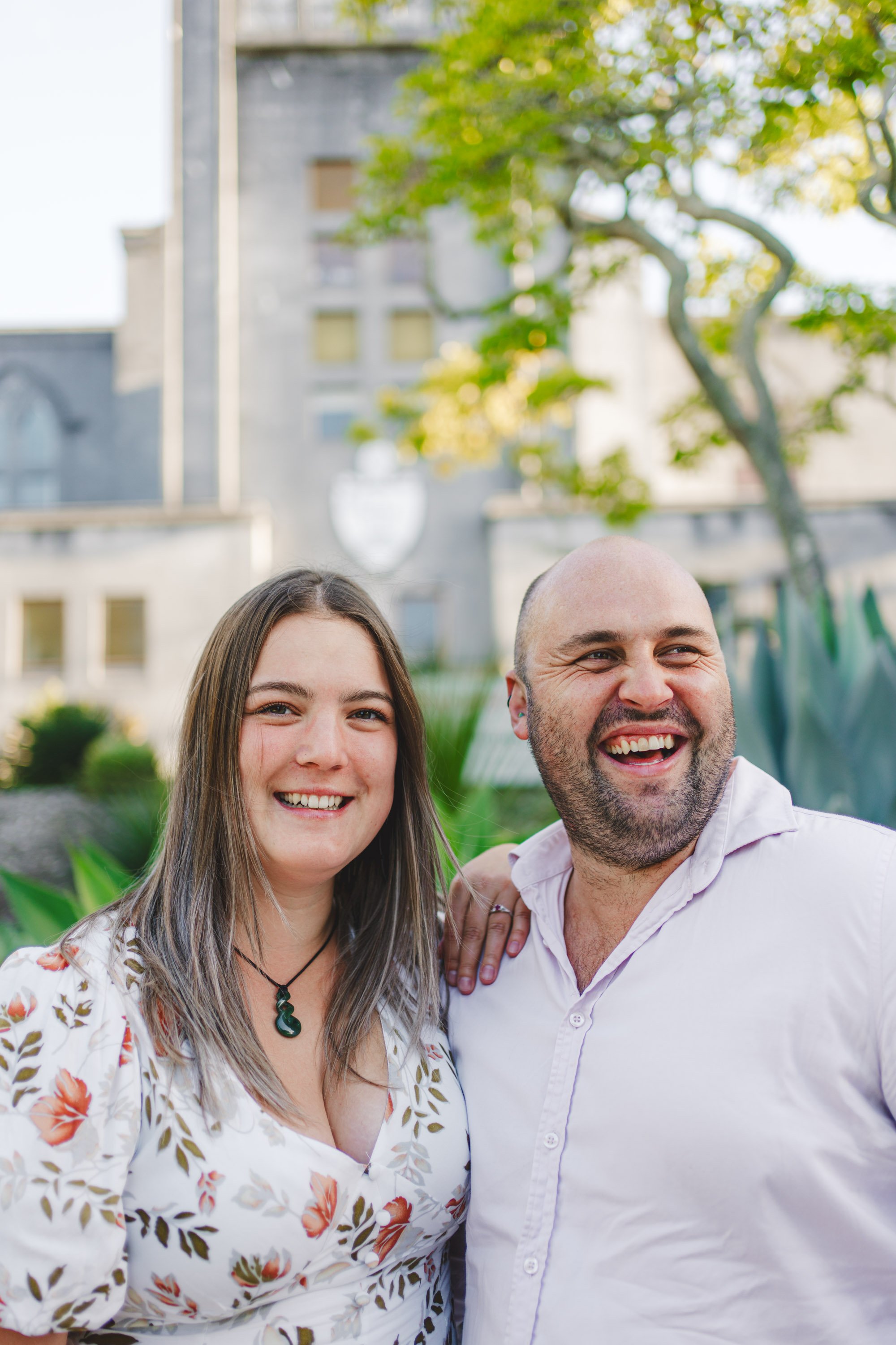 A smiling woman with long brown hair and a man with a bald head, beard, and mustache, standing outdoors in front of green plants and a building, with the woman resting her hand on the man's shoulder.