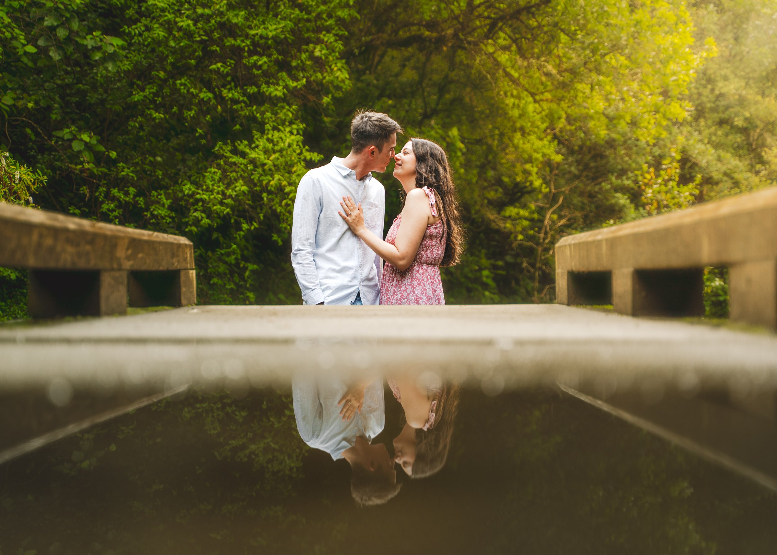 A young couple standing closely on a bridge surrounded by lush green trees, with their reflection visible in water below.