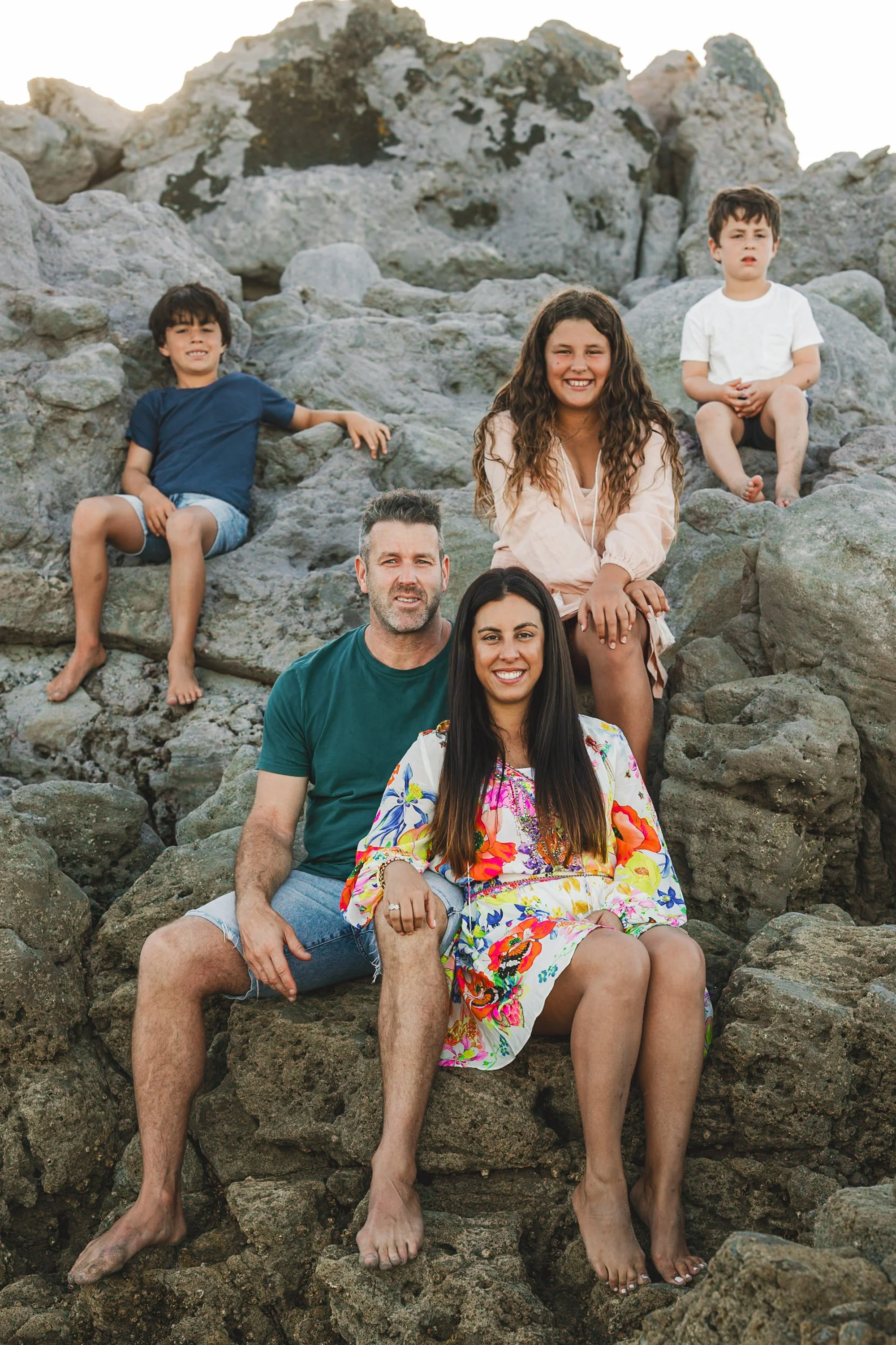 A family of five sitting on rocks near the beach, smiling and enjoying the outdoors.