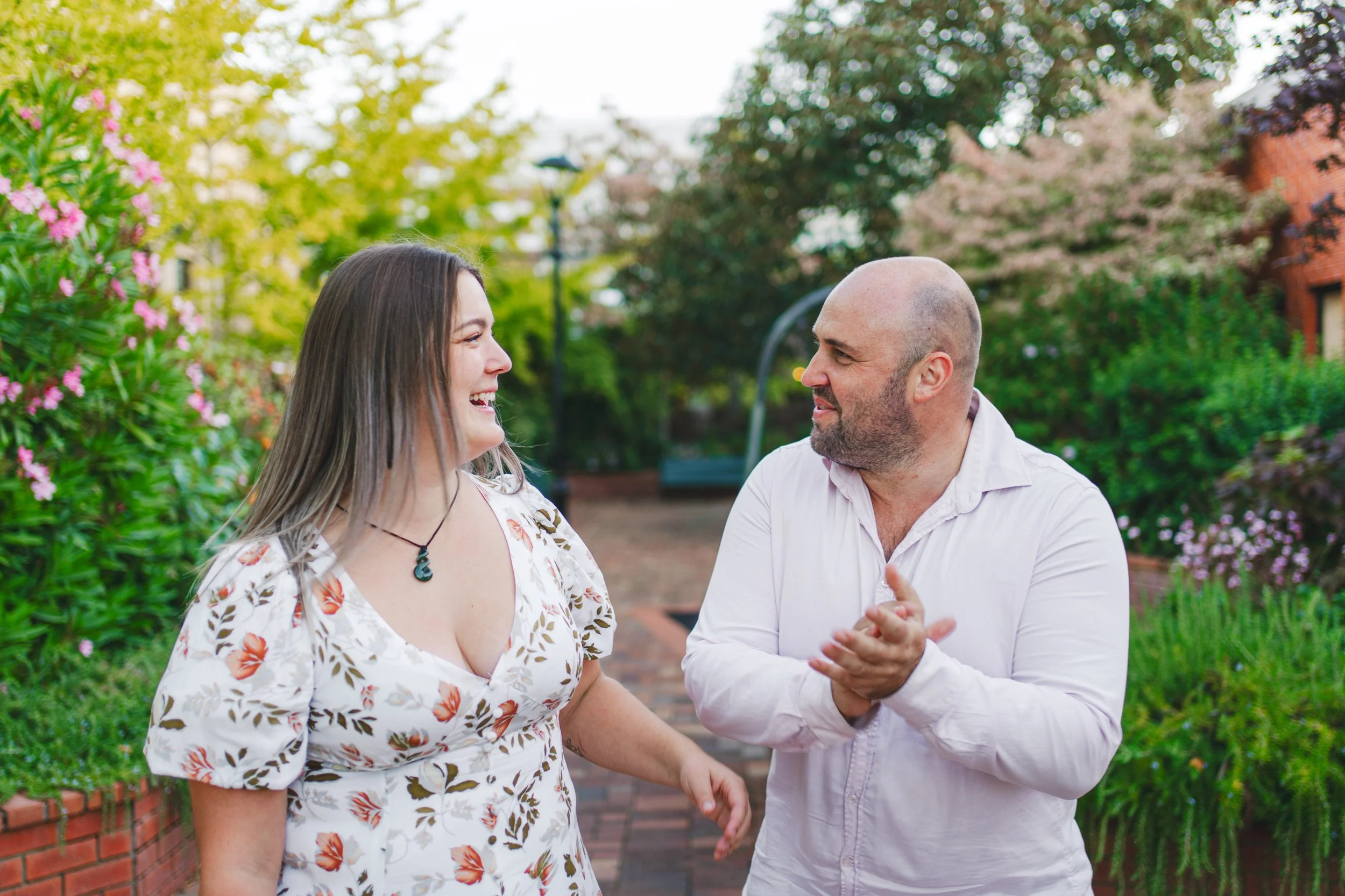 A man and woman smiling and laughing while talking outdoors in a garden with greenery and flowers.