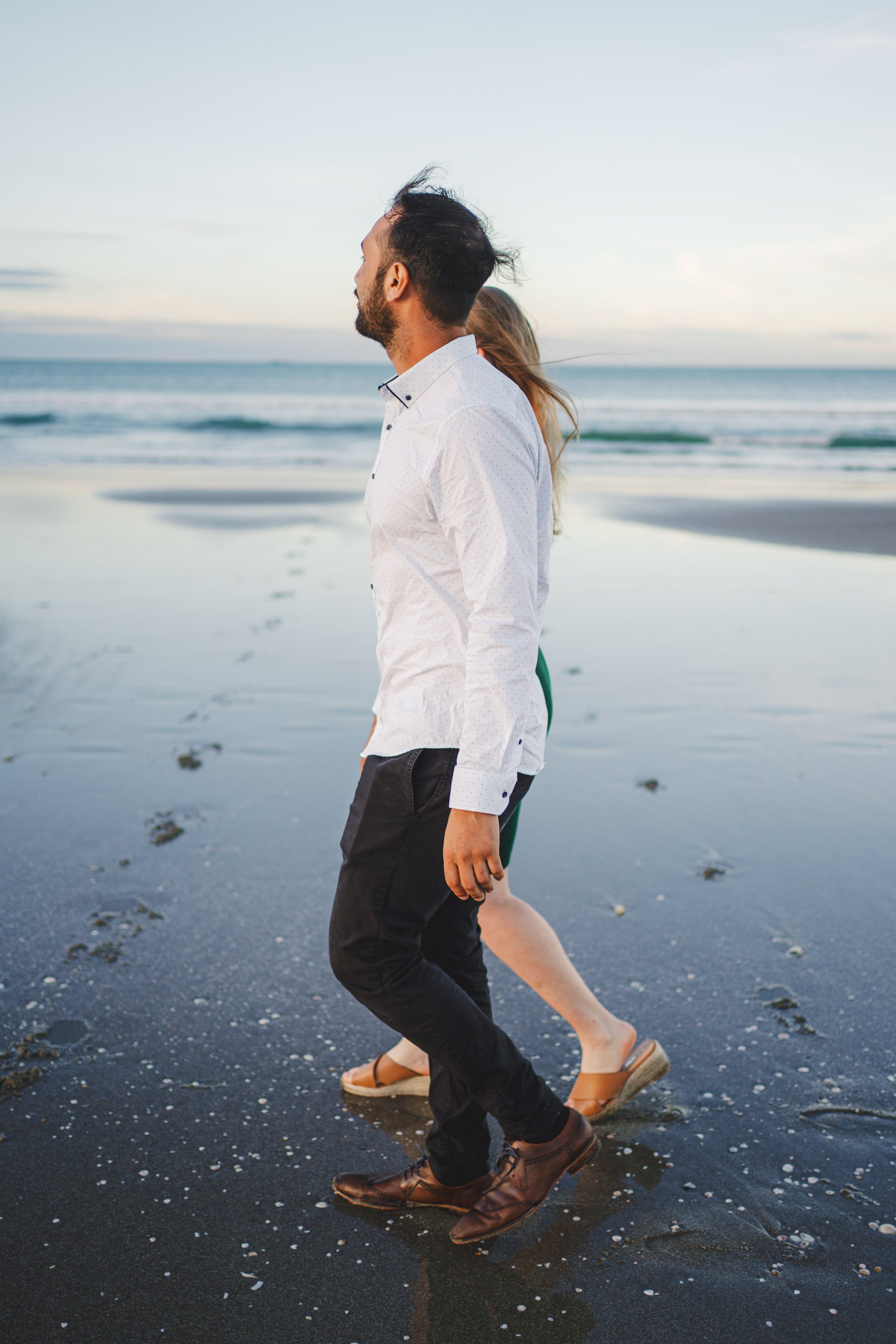 A man and woman walking on the beach at sunset, with the man in the foreground wearing a white shirt and black pants, and the woman behind him in sandals, partly obscured.