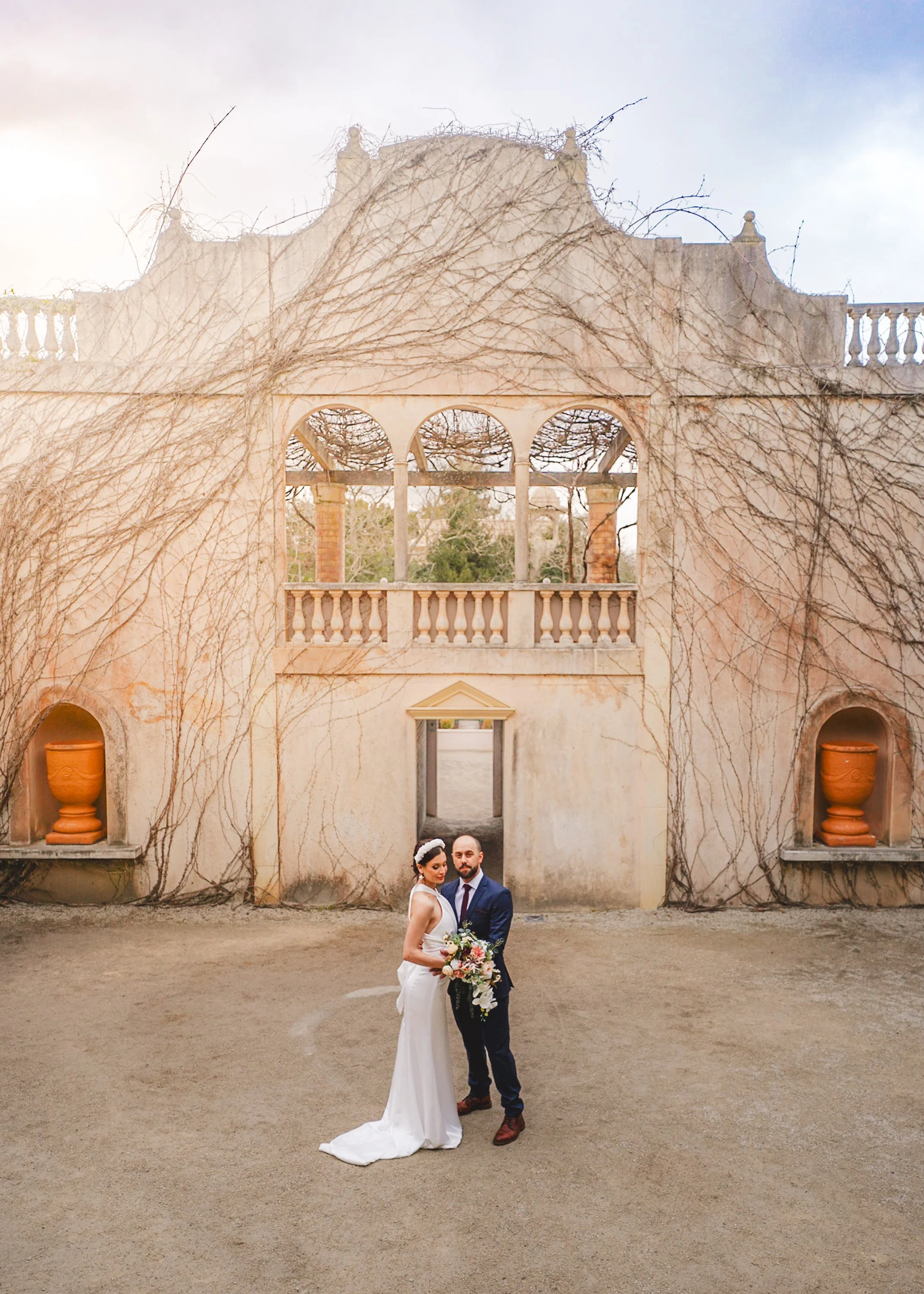 A bride and groom standing together in wedding attire in front of a historic building with vine-covered walls, columns, and terracotta planters.
