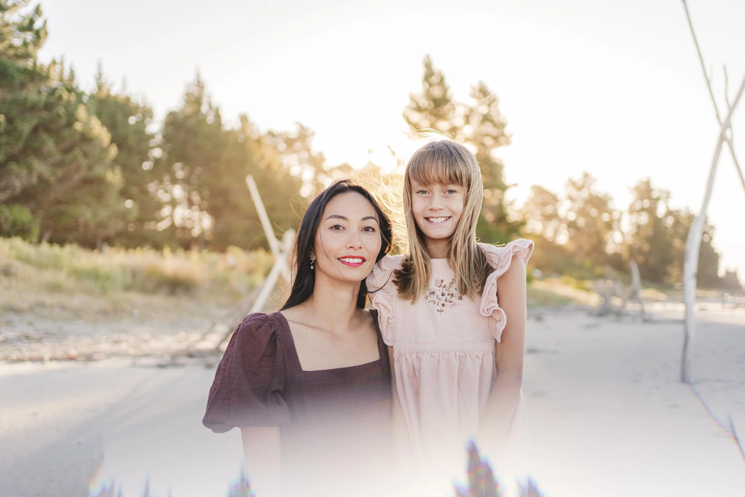 A woman and a young girl smiling on the beach during sunset, with trees in the background.