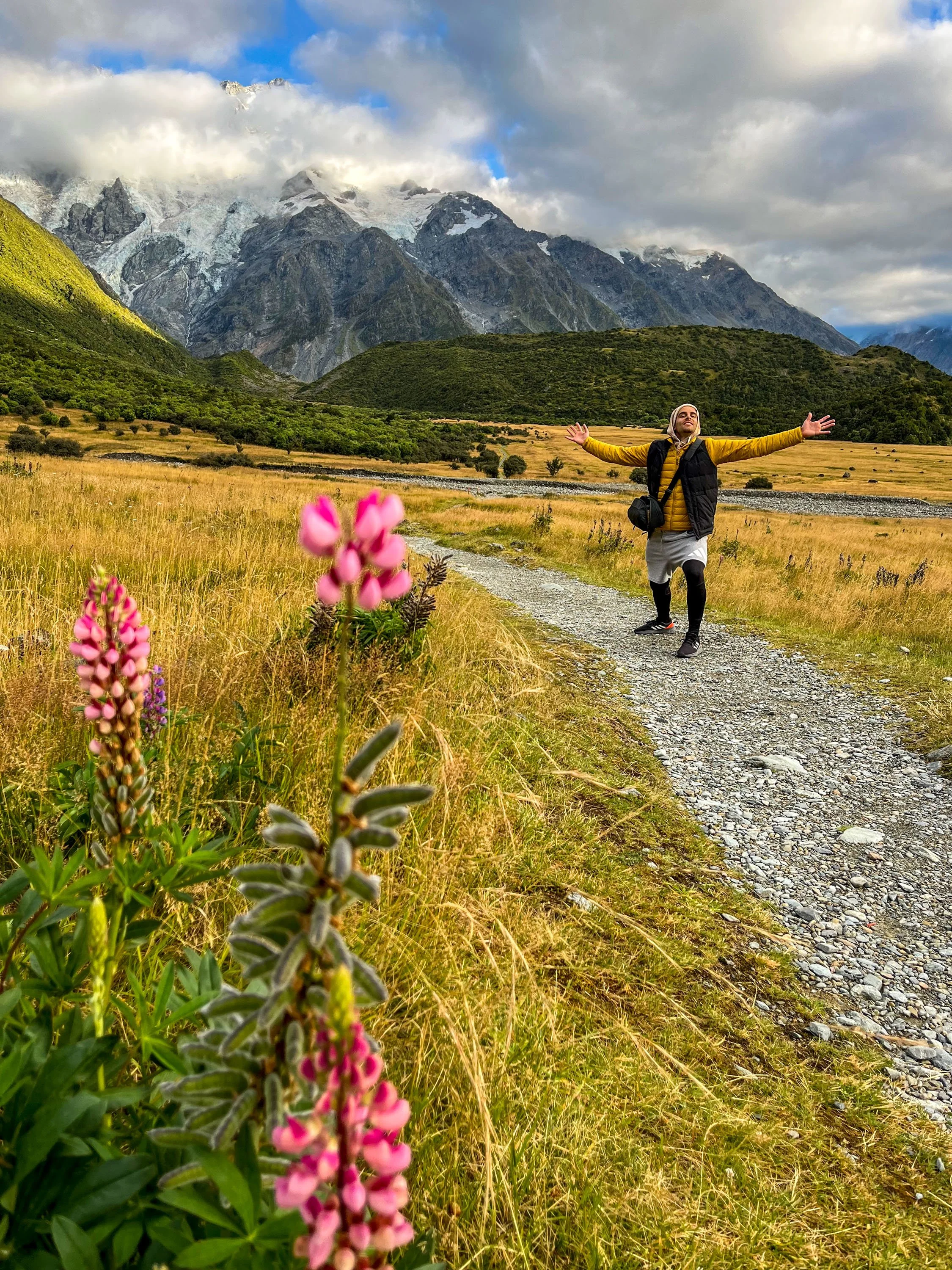 A person standing on a gravel trail in a field with yellowish grass, arms wide open, with mountains in the background, and pink flowers in the foreground.