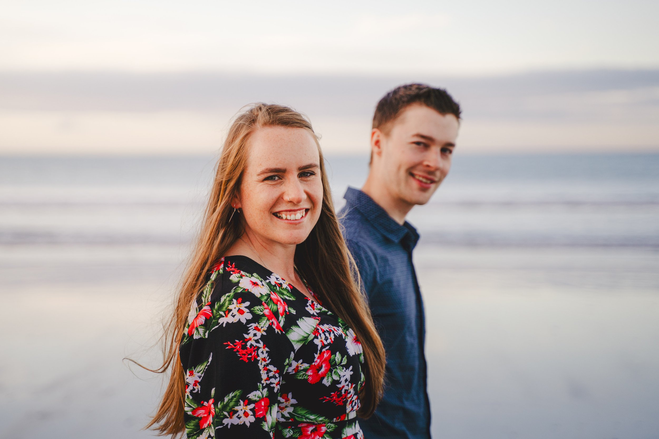 A smiling young woman with long red hair and a floral dress stands on a beach with a young man in a blue shirt behind her, with the ocean and sky in the background.