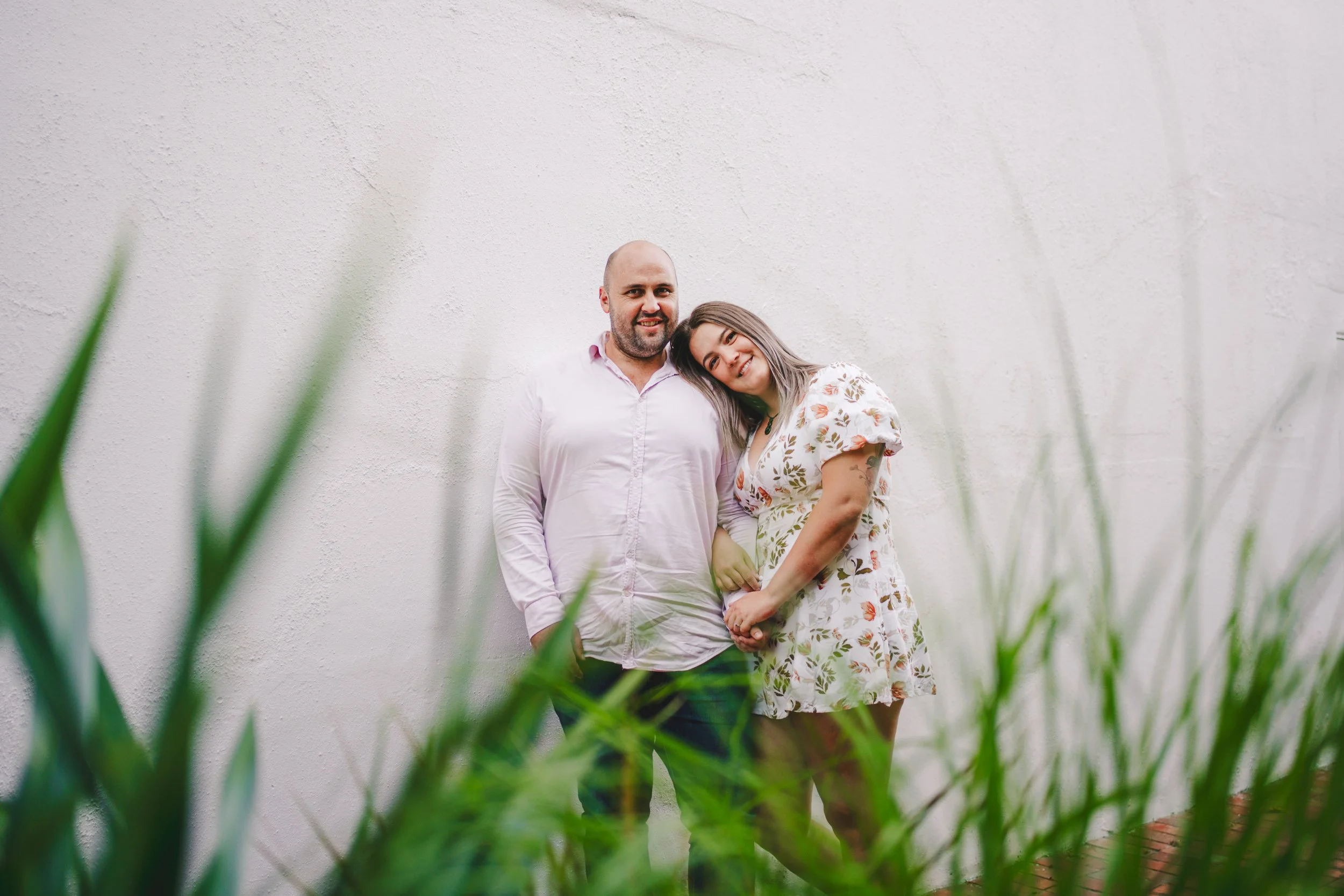A couple standing close together outdoors, smiling, against a white textured wall with green plants in the foreground.