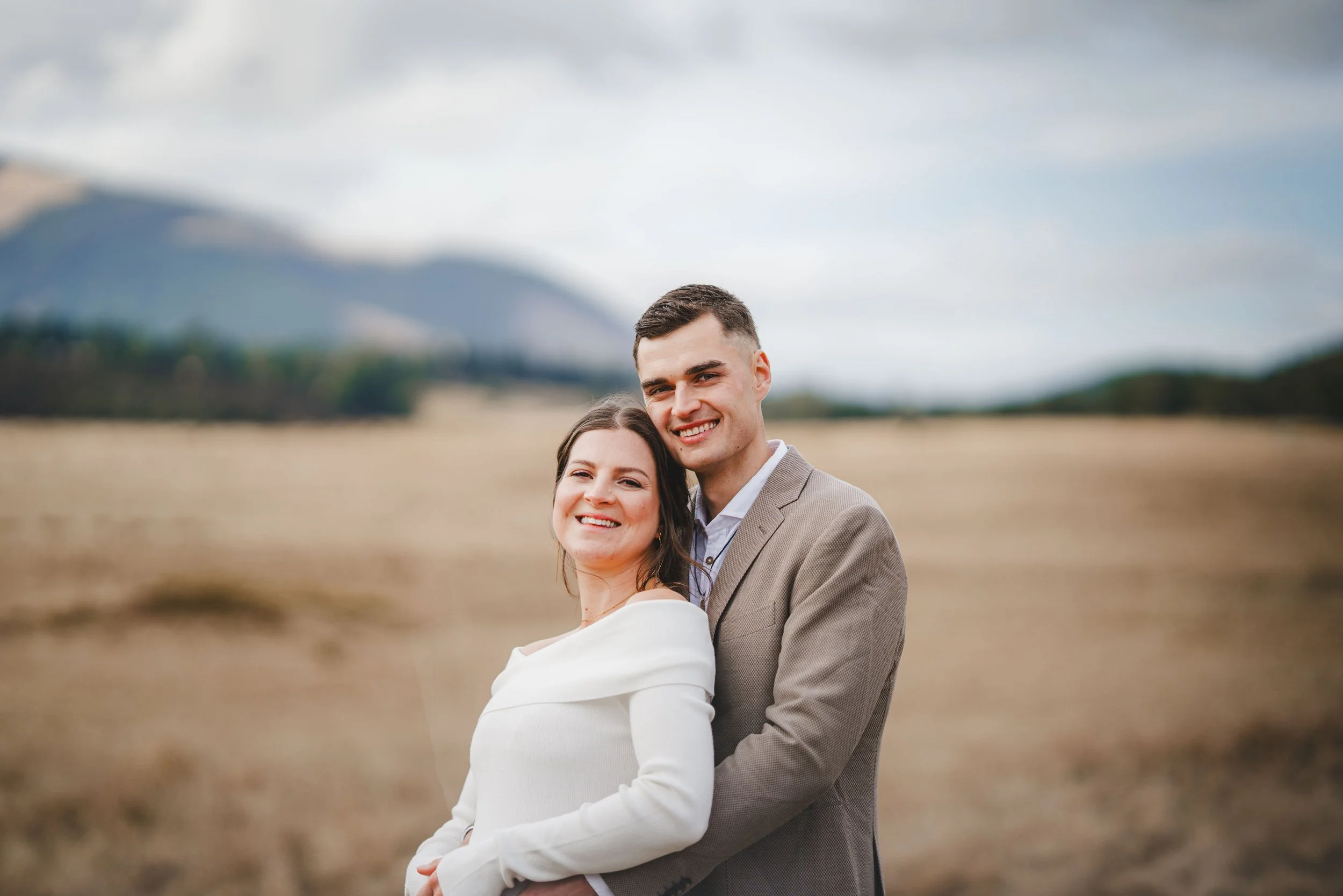A smiling couple standing close together outdoors with a mountain landscape in the background.