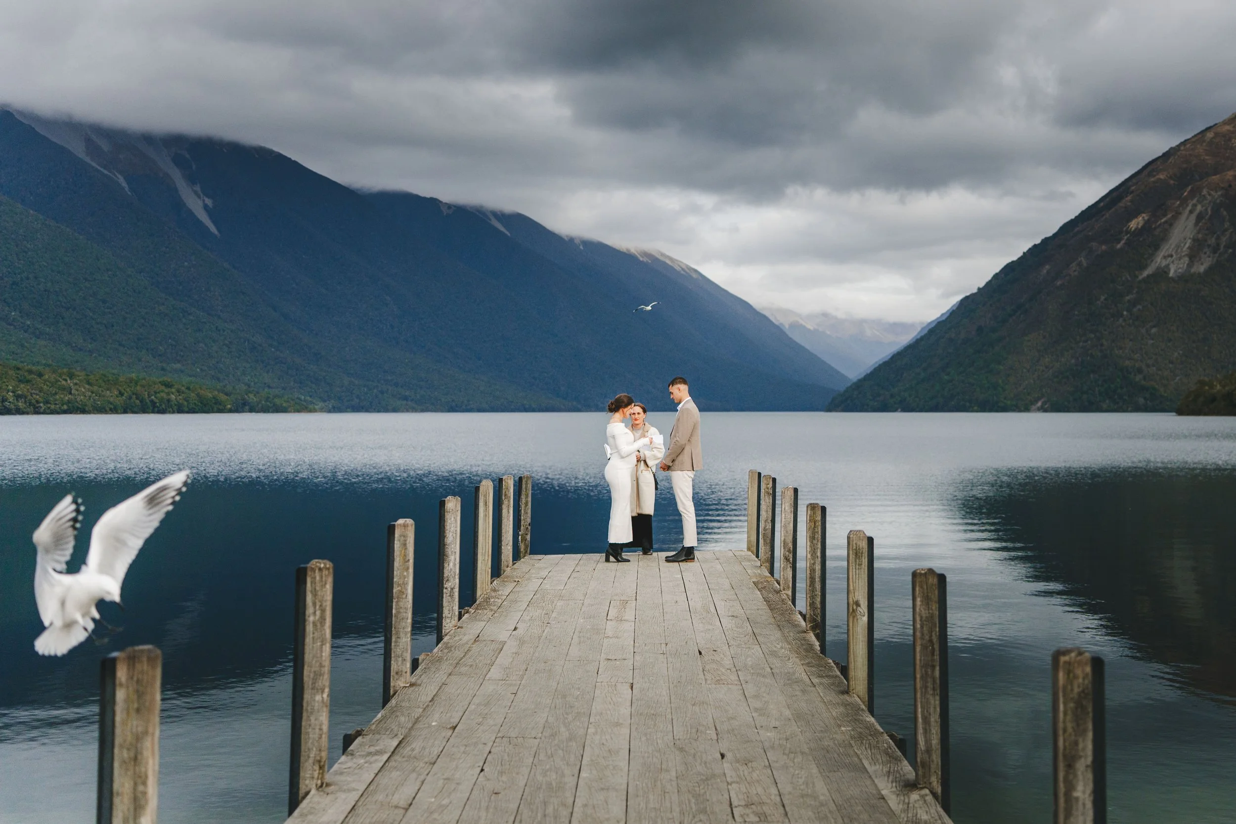 A couple getting married standing on a wooden dock over a calm lake, with mountains and cloudy sky in the background, and birds flying nearby.