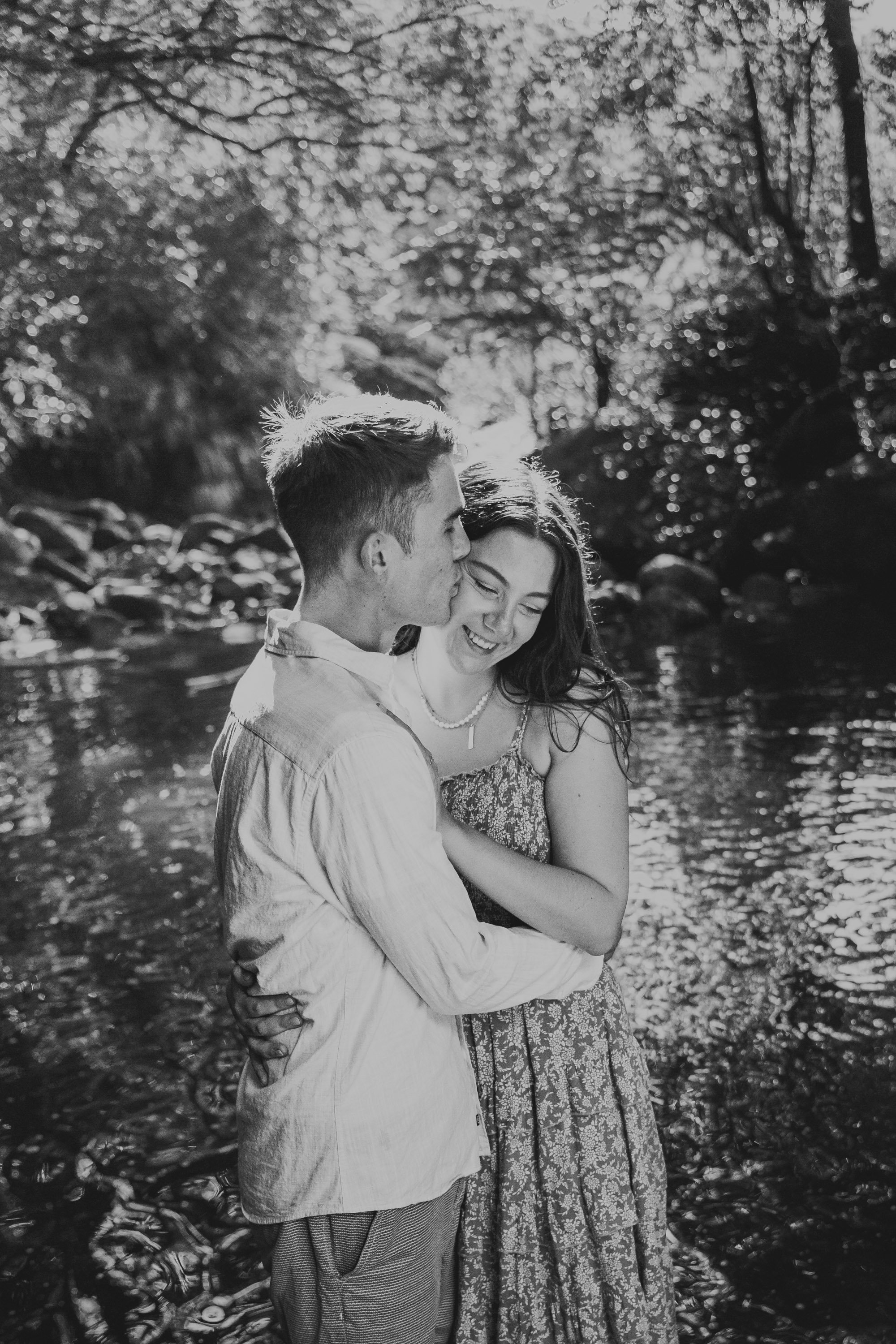 A young couple is embracing and smiling by a river in a forest, with sunlight filtering through the trees.