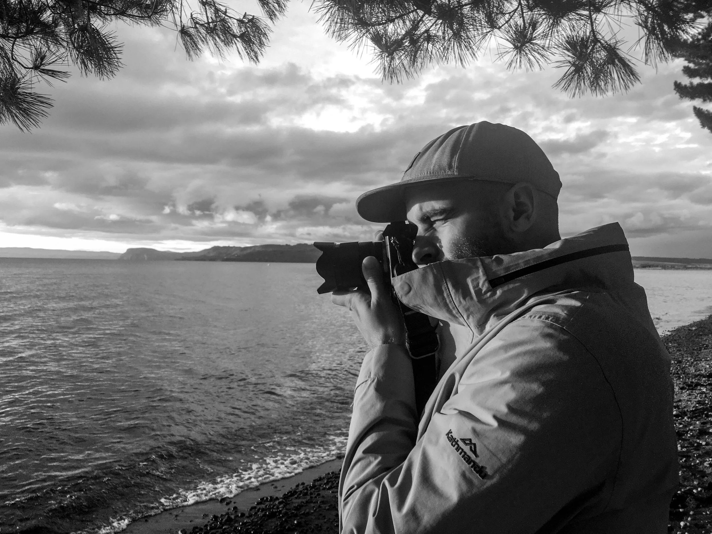 A man wearing a cap and a windbreaker jacket is taking a photograph by the water's edge with a camera, with clouds and mountains in the background, in black and white.