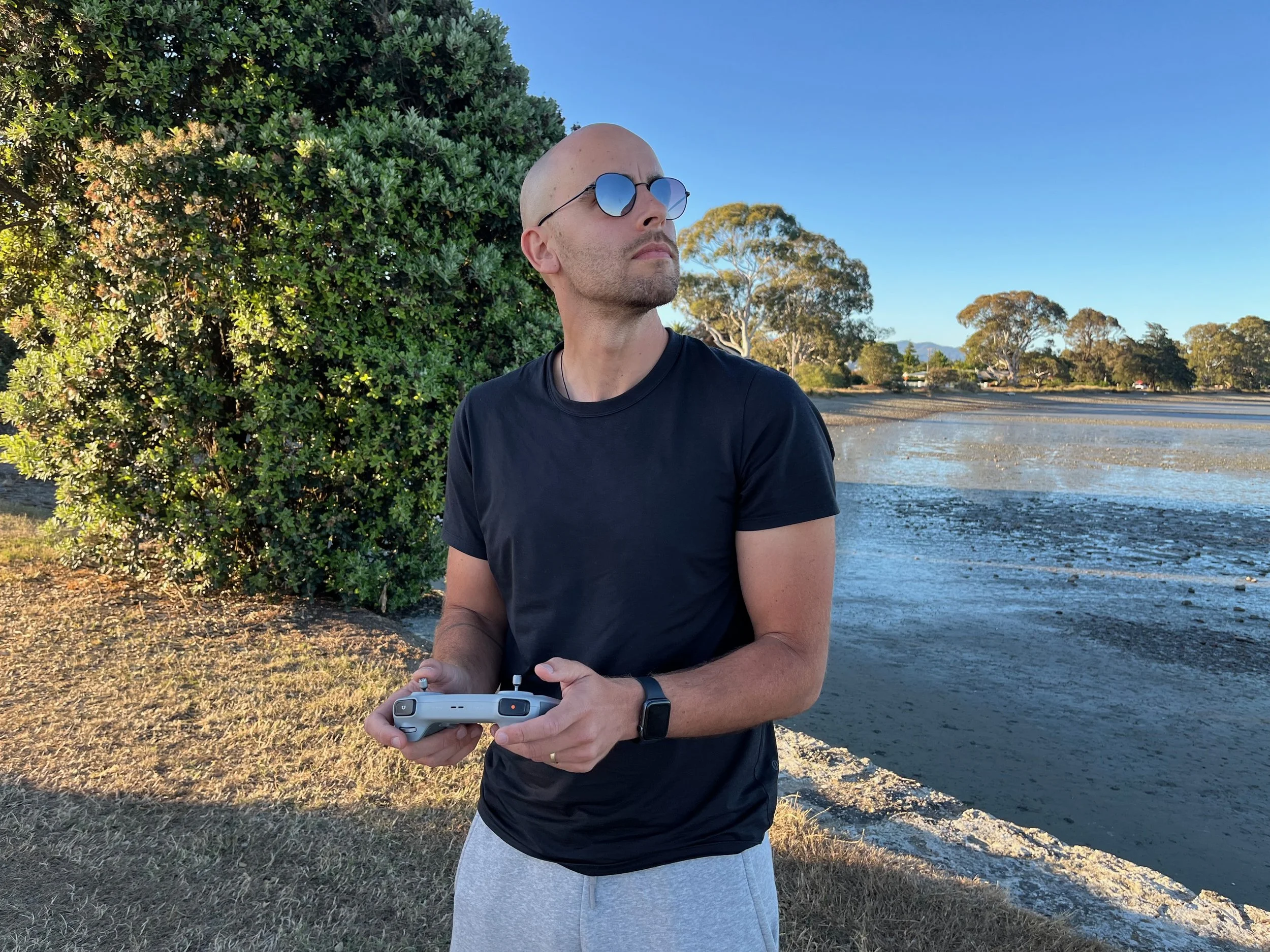 A man with a shaved head wearing sunglasses, a black T-shirt, and light gray shorts standing outdoors near water and trees, holding a drone remote controller.