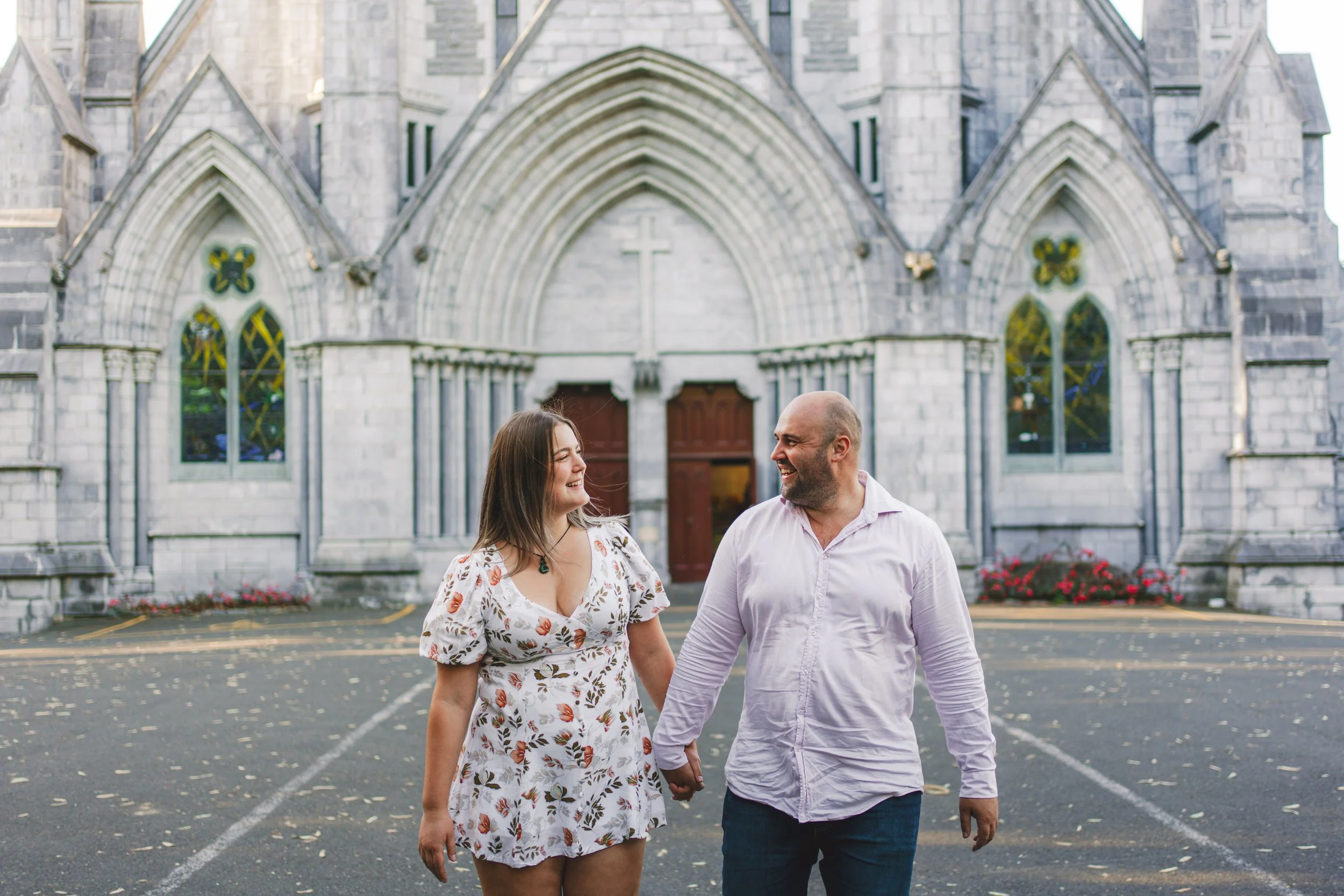 A couple holding hands and smiling at each other in front of a Gothic-style church.