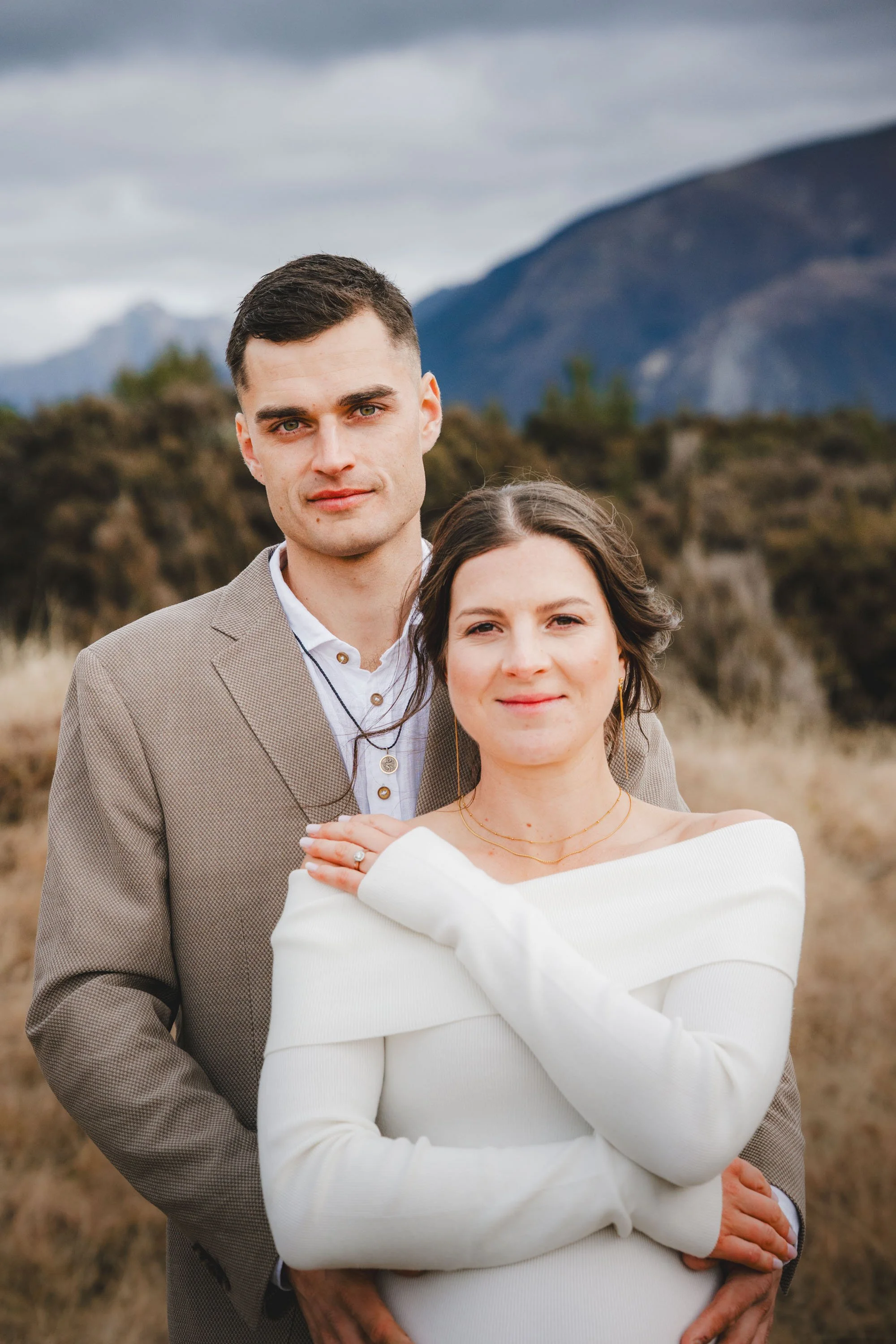 A young couple outdoors in a mountainous area, standing close together, with the woman in a white off-shoulder top and the man in a light brown blazer. The woman has shoulder-length brown hair, and the man has short dark hair. Overcast sky and mounta