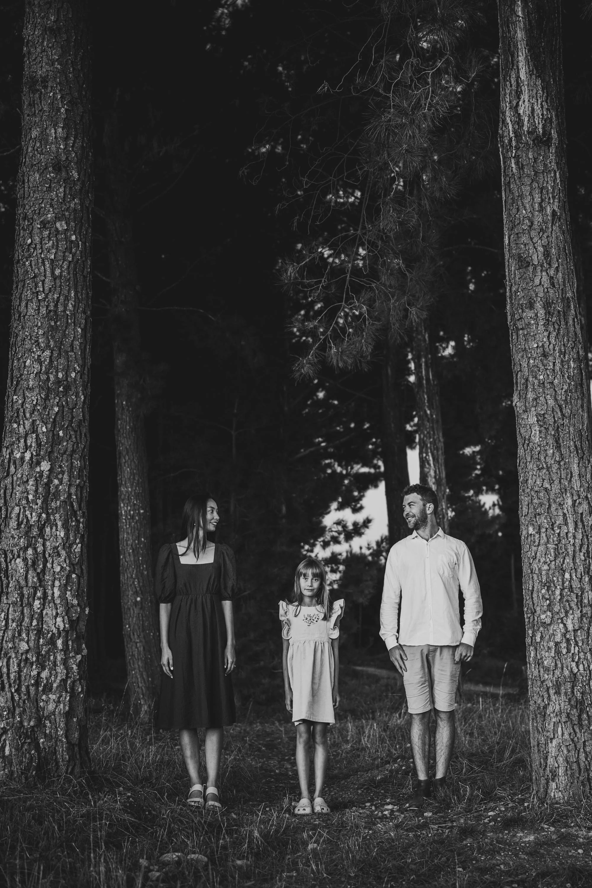 A black and white photo of three people, a woman, a young girl, and a man, walking through a wooded forest area surrounded by tall trees.
