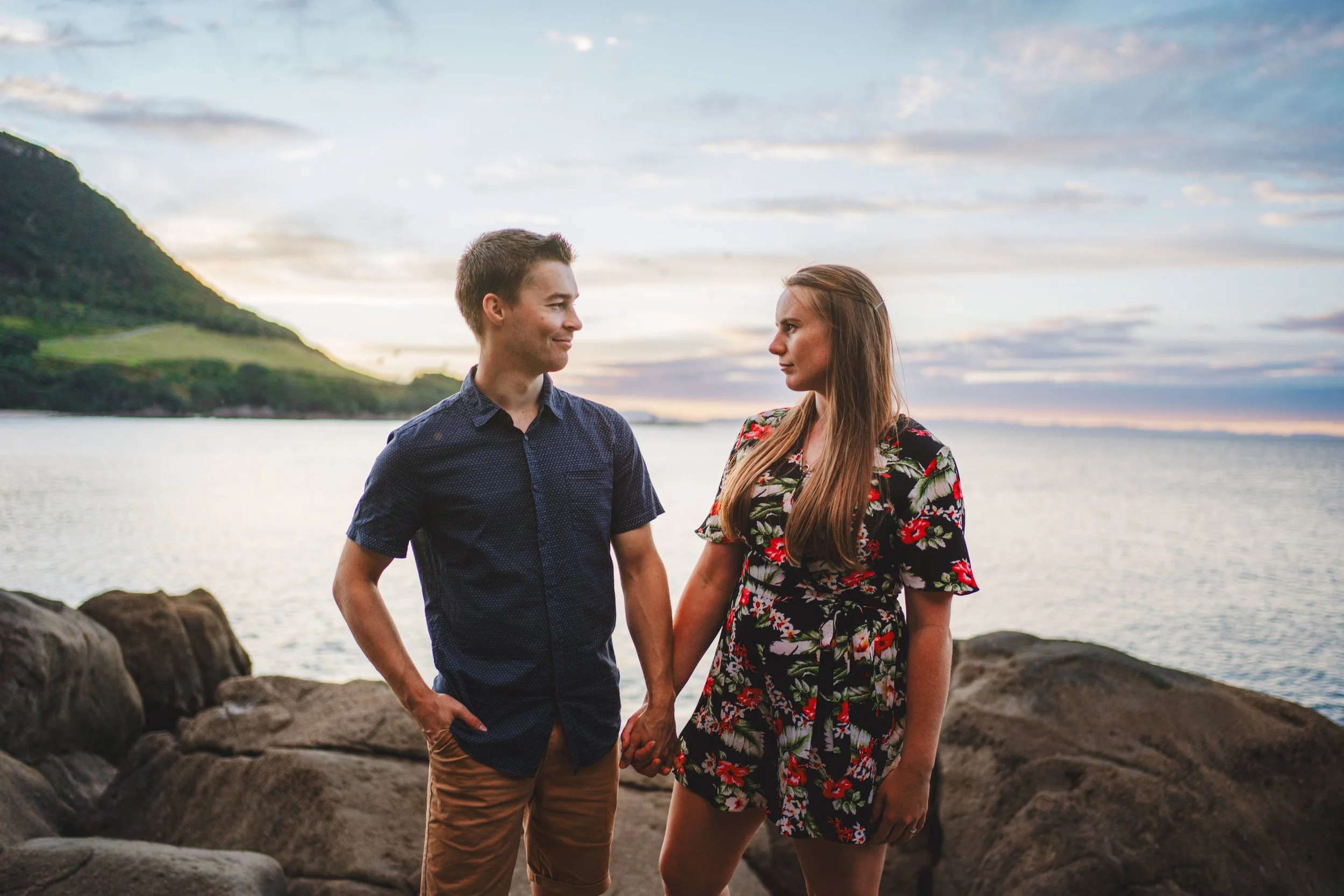 A young man and woman holding hands on rocks by a lake at sunset, looking at each other.