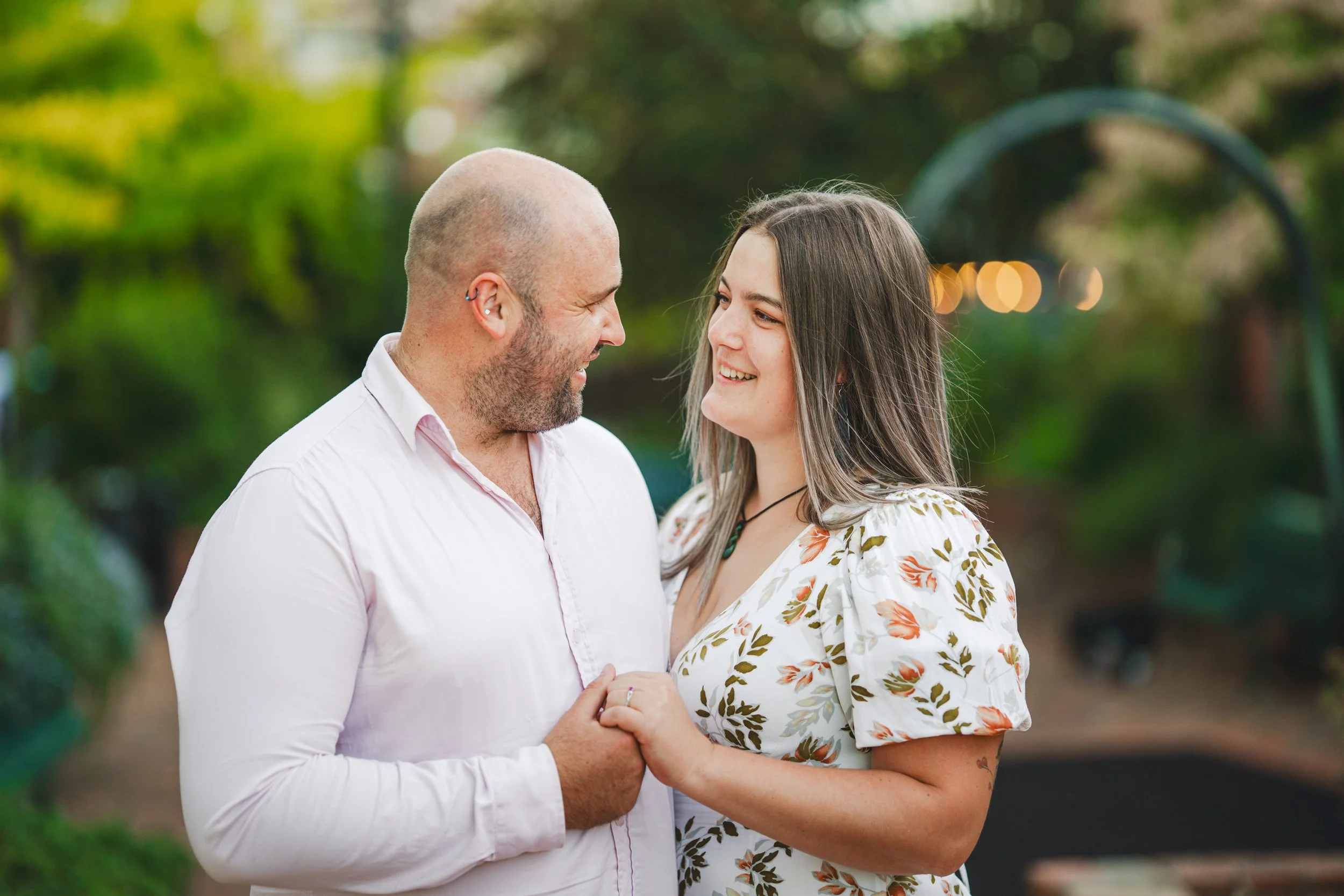 A couple holding hands and smiling at each other outdoors in a lush, green garden with blurred background lighting.