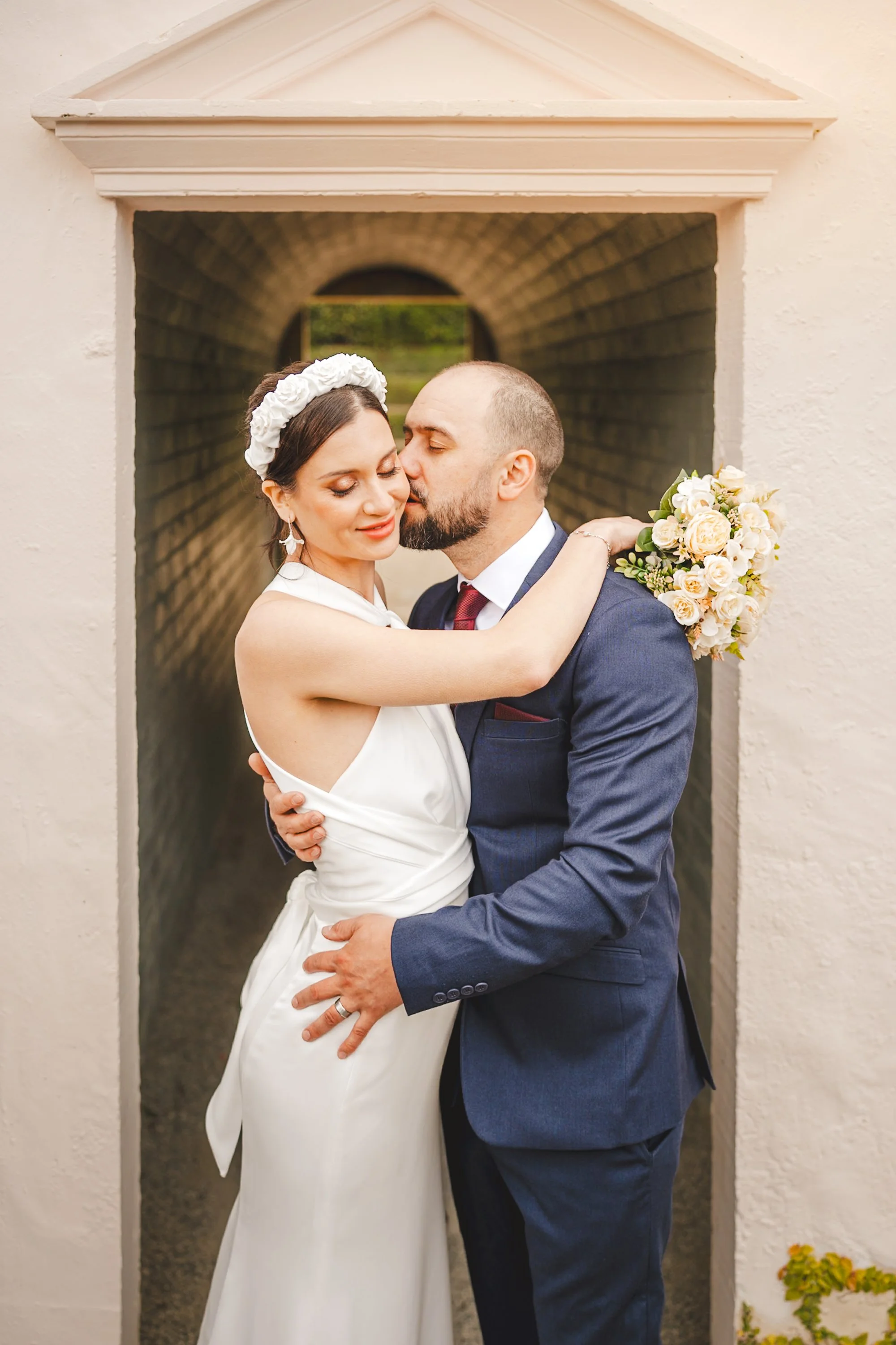 A bride and groom embrace in front of a small archway. The bride wears a white wedding dress and a floral headband, holding a bouquet of white and cream roses. The groom wears a navy suit and a maroon tie. They are sharing a tender moment, with the g