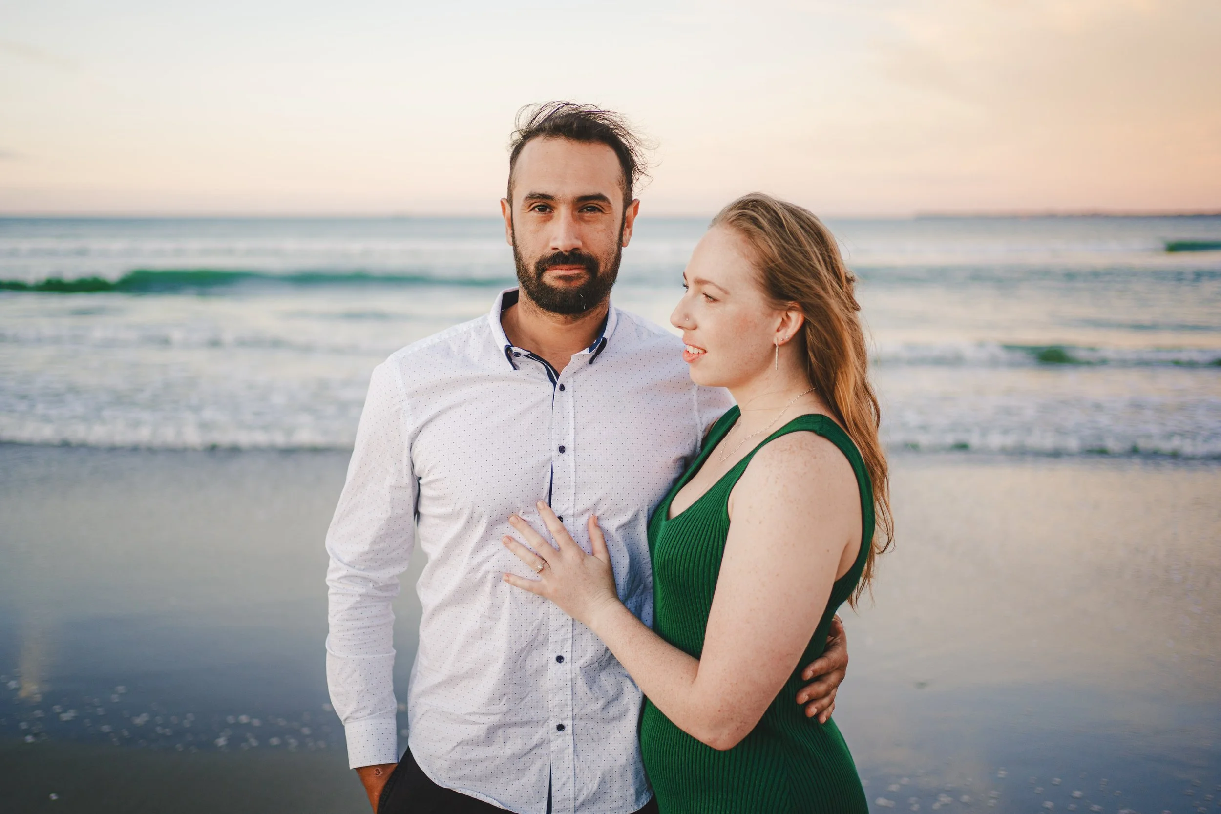 A man and woman stand close together on a beach with ocean waves in the background, at sunset.