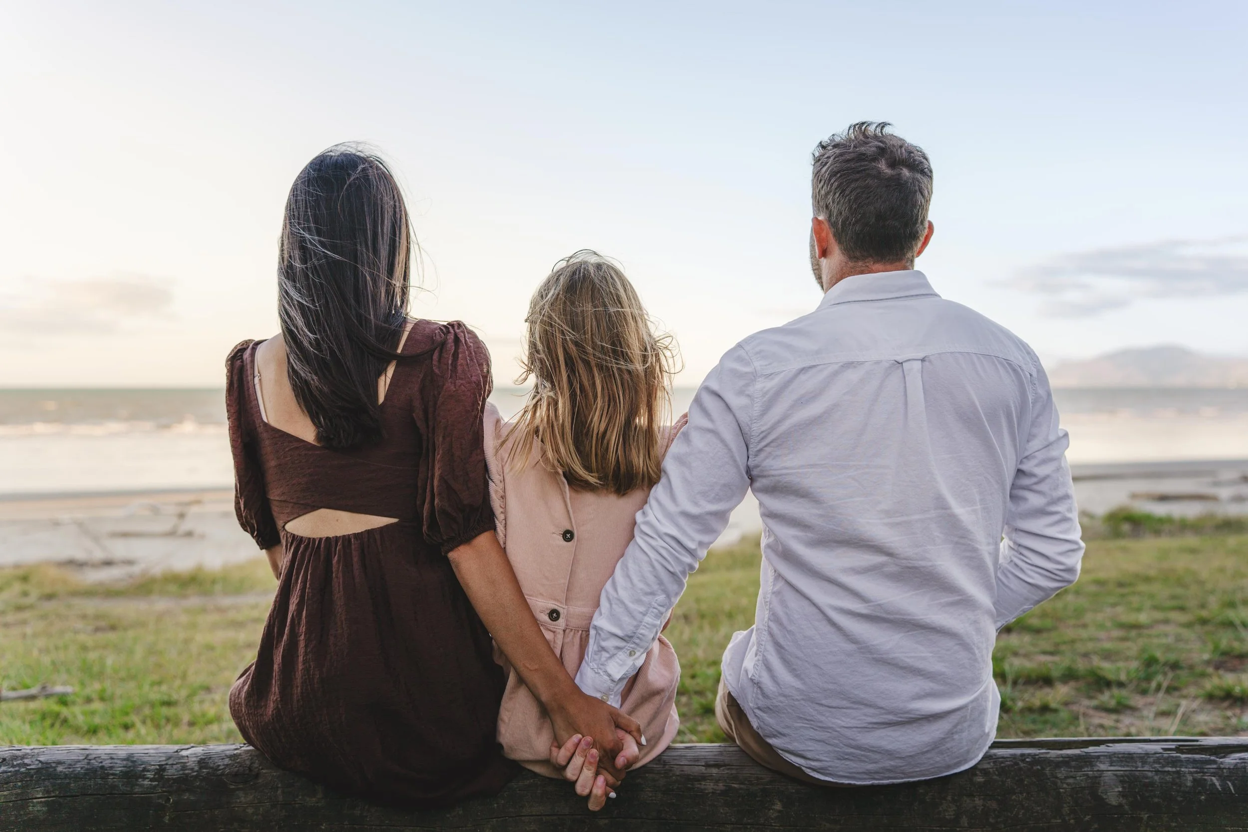 A family of three sitting on a log at the beach, holding hands and facing the ocean under a cloudy sky.
