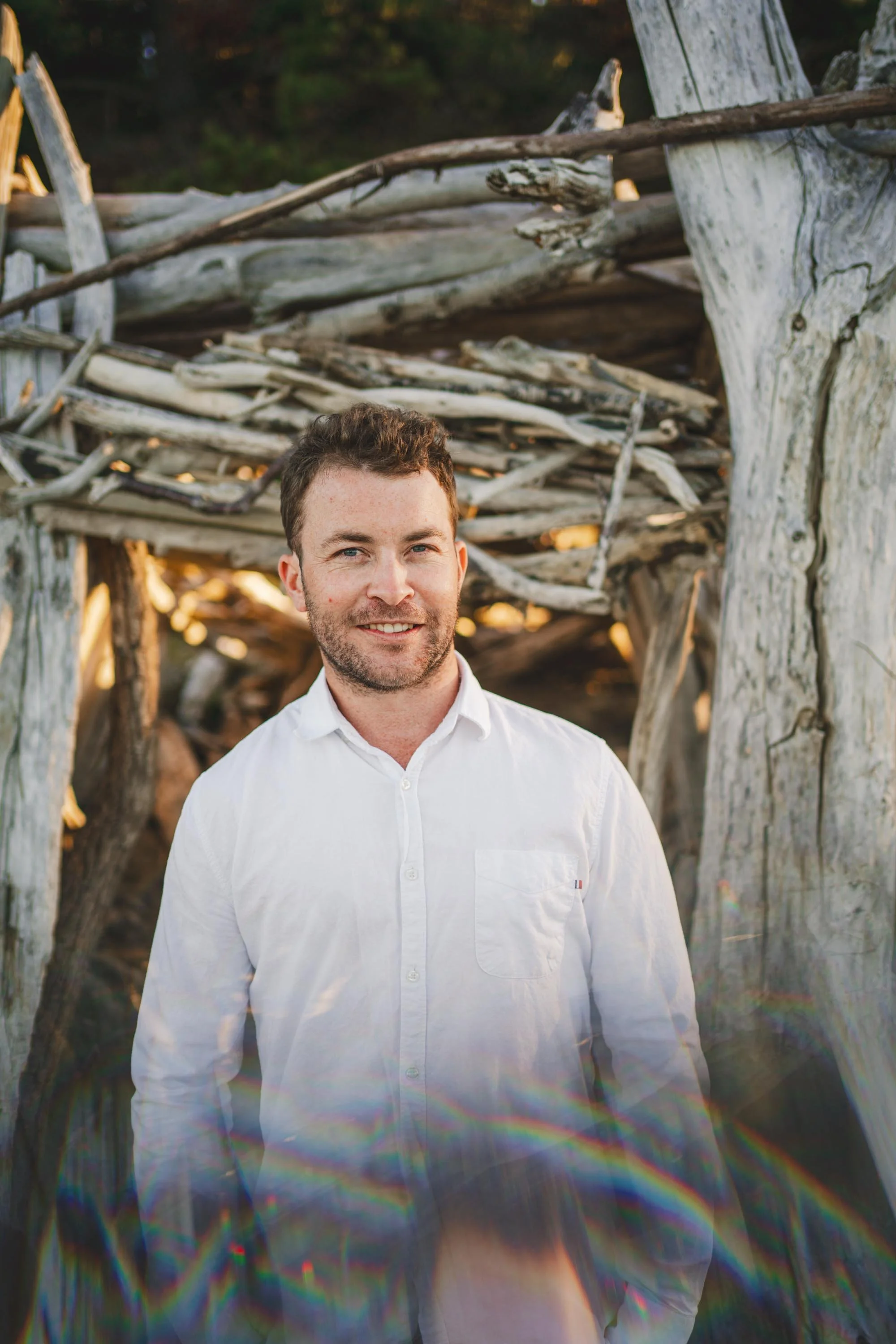 A man in a white shirt standing outdoors in front of a driftwood structure, with rainbow light refractions visible at the bottom of the image.