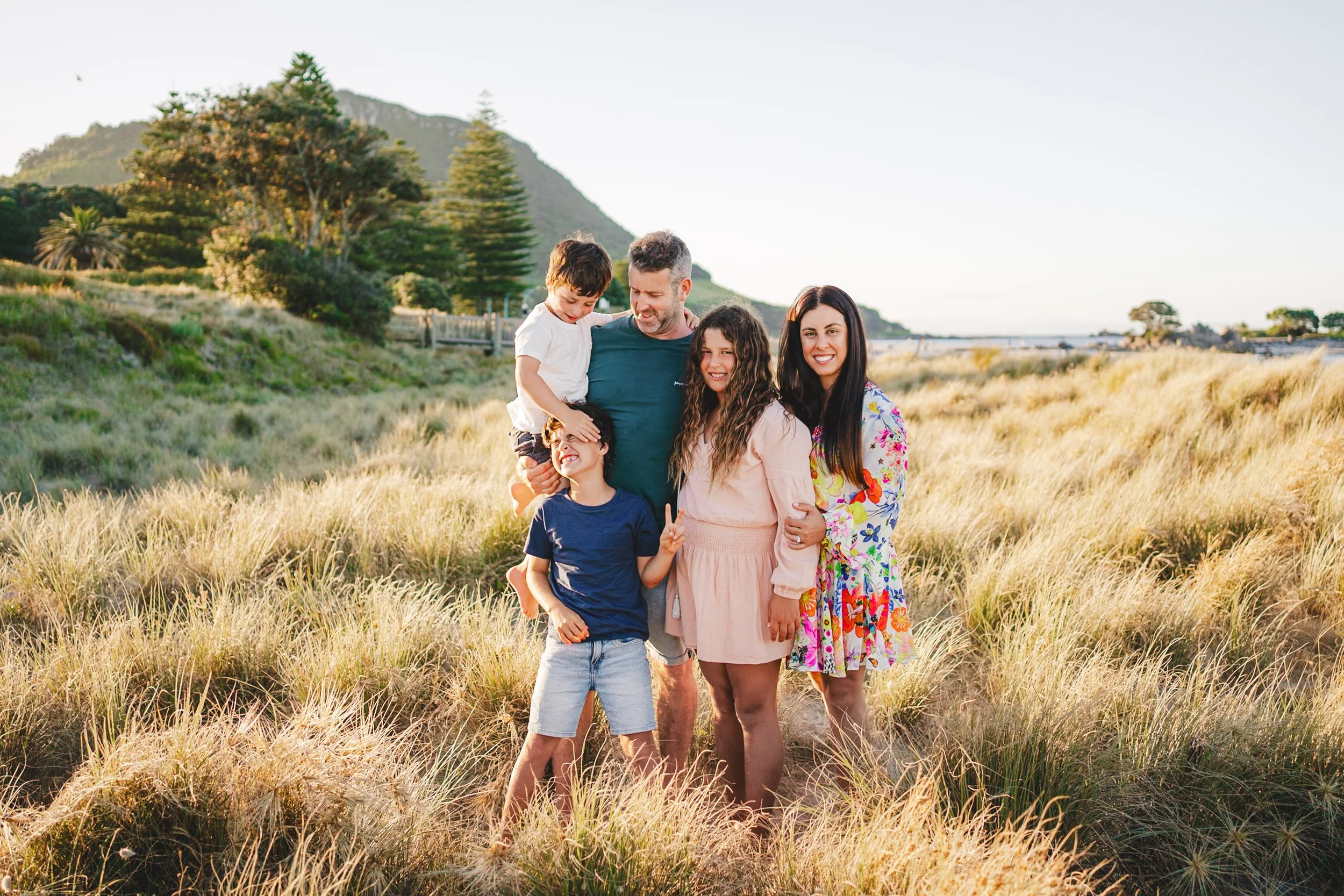 Family of six standing in a grassy field with green hills and trees in the background, during sunset or late afternoon.