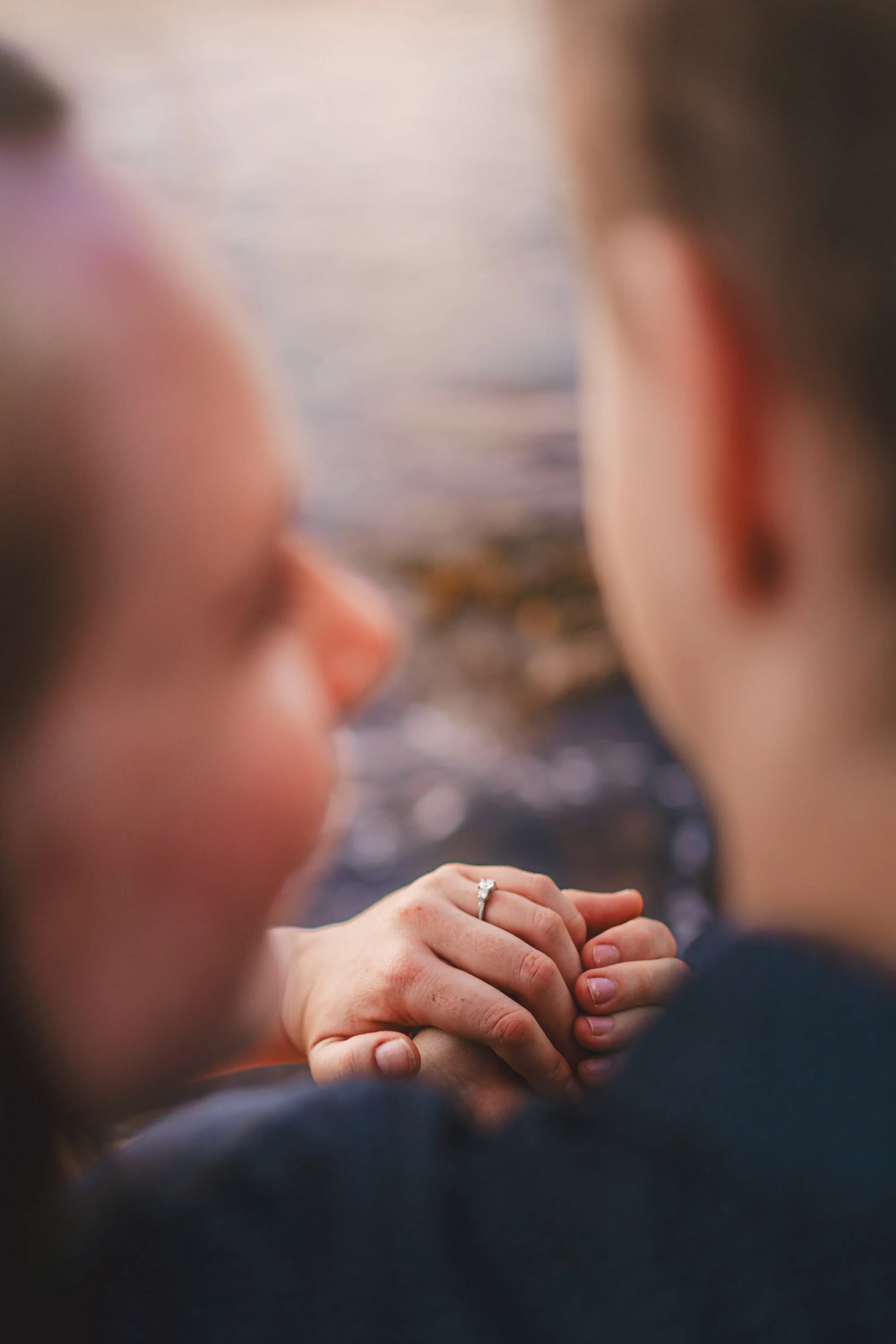 Close-up of a couple holding hands with an engagement ring, near a body of water at sunset, with two women in the foreground blurred.