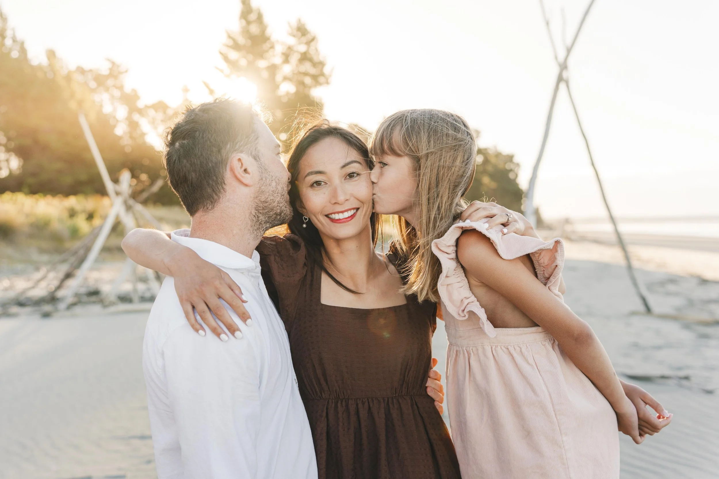 Family of three at the beach during sunset, with the mother smiling as both parents kiss her on the cheeks, and the daughter giving her a kiss on the cheek.