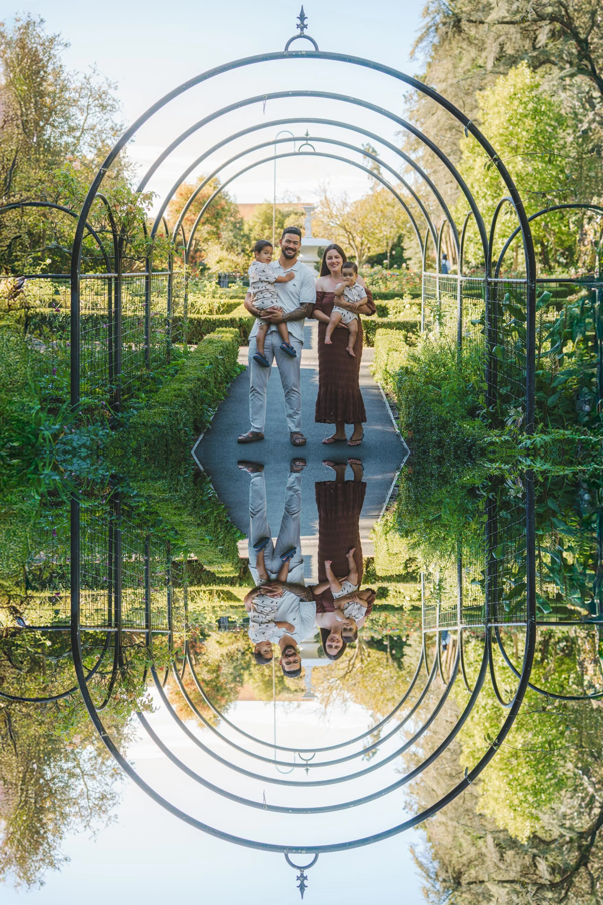 Family of four, two adults and two children, standing in a garden with an archway overhead and a reflective surface on the ground creating a mirror effect.
