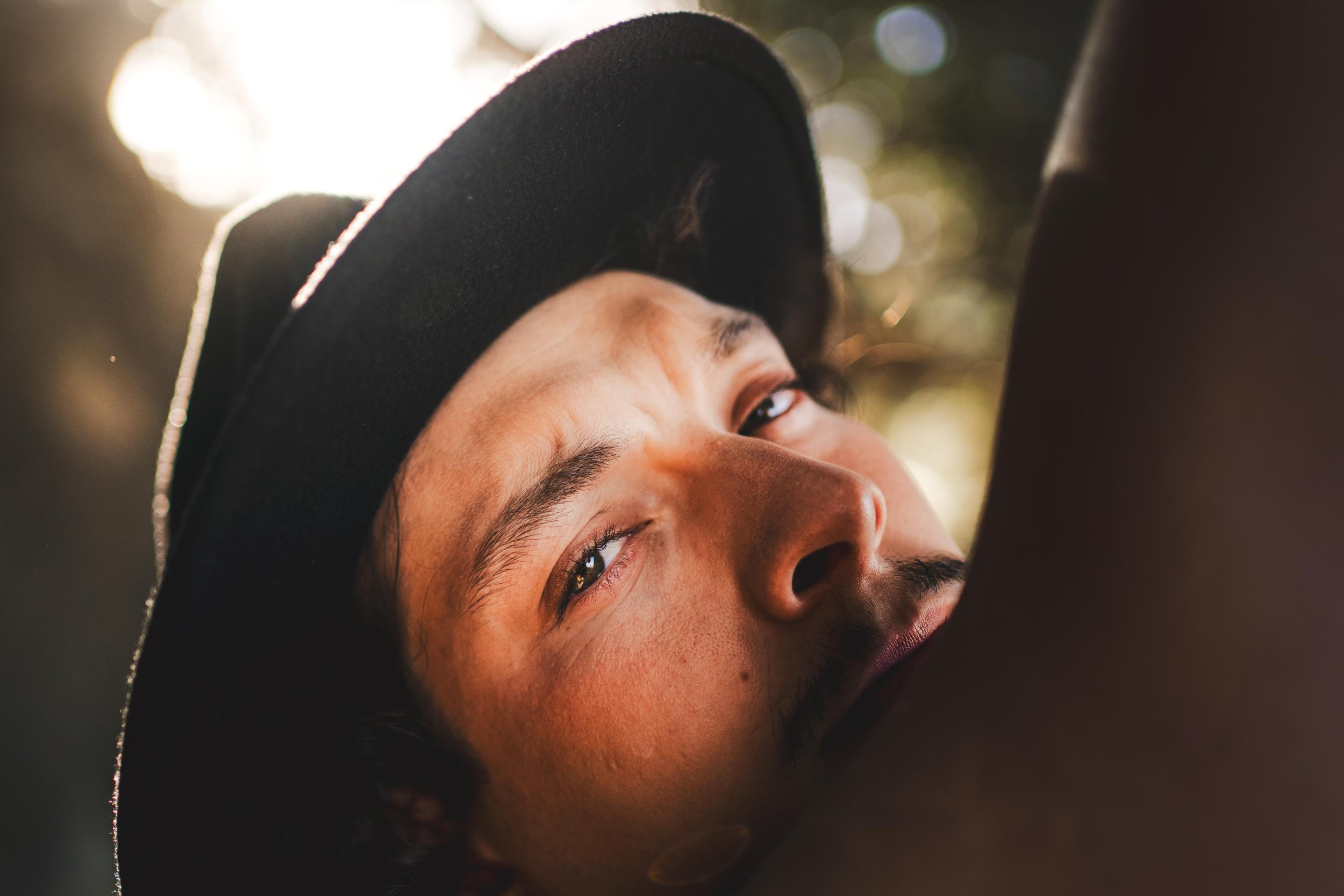 A close-up photo of a man with a relaxed expression, wearing a black hat and looking into the camera, with sunlight in the background.