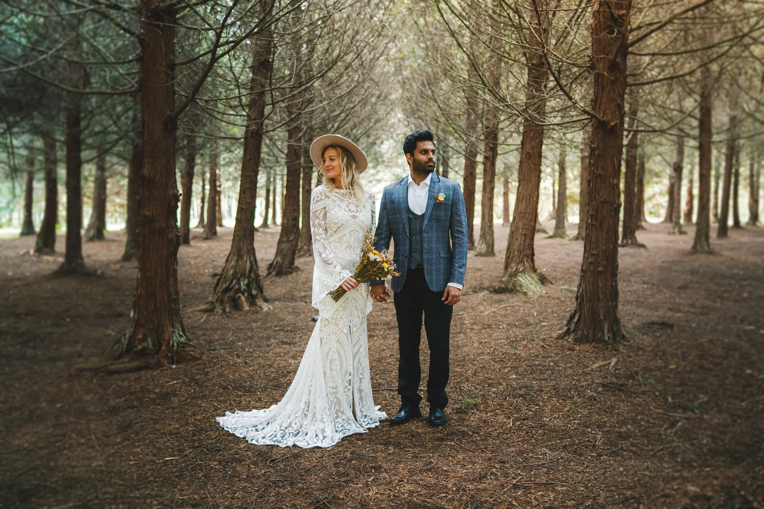 A bride and groom stand hand in hand in a forest, the bride in a white lace wedding dress, holding a bouquet, and the groom in a blue checked suit with a white shirt and boutonniere.