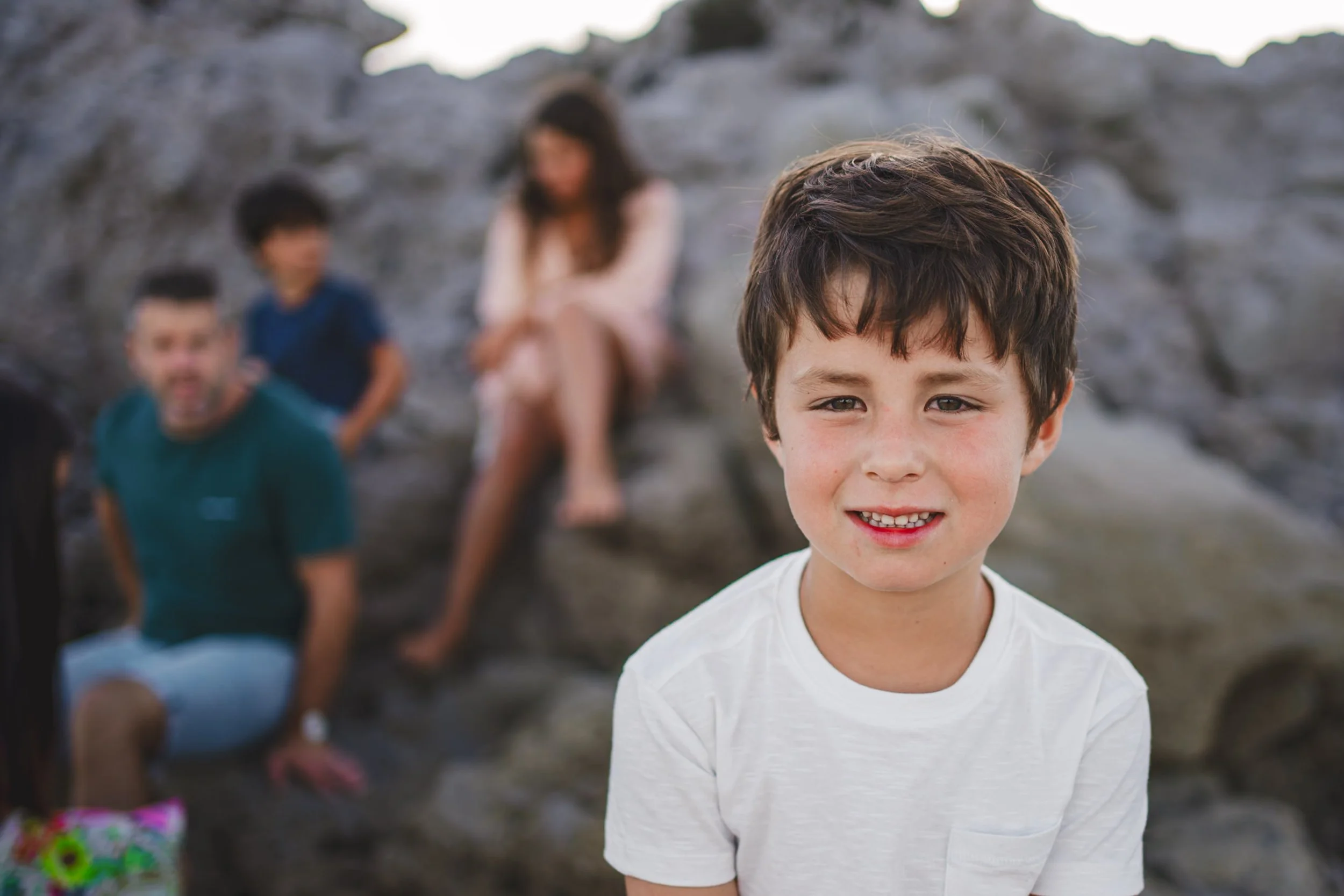 Young boy with brown hair and a white shirt smiling in front of a group of people sitting on rocks, with a rocky outdoor setting in the background.