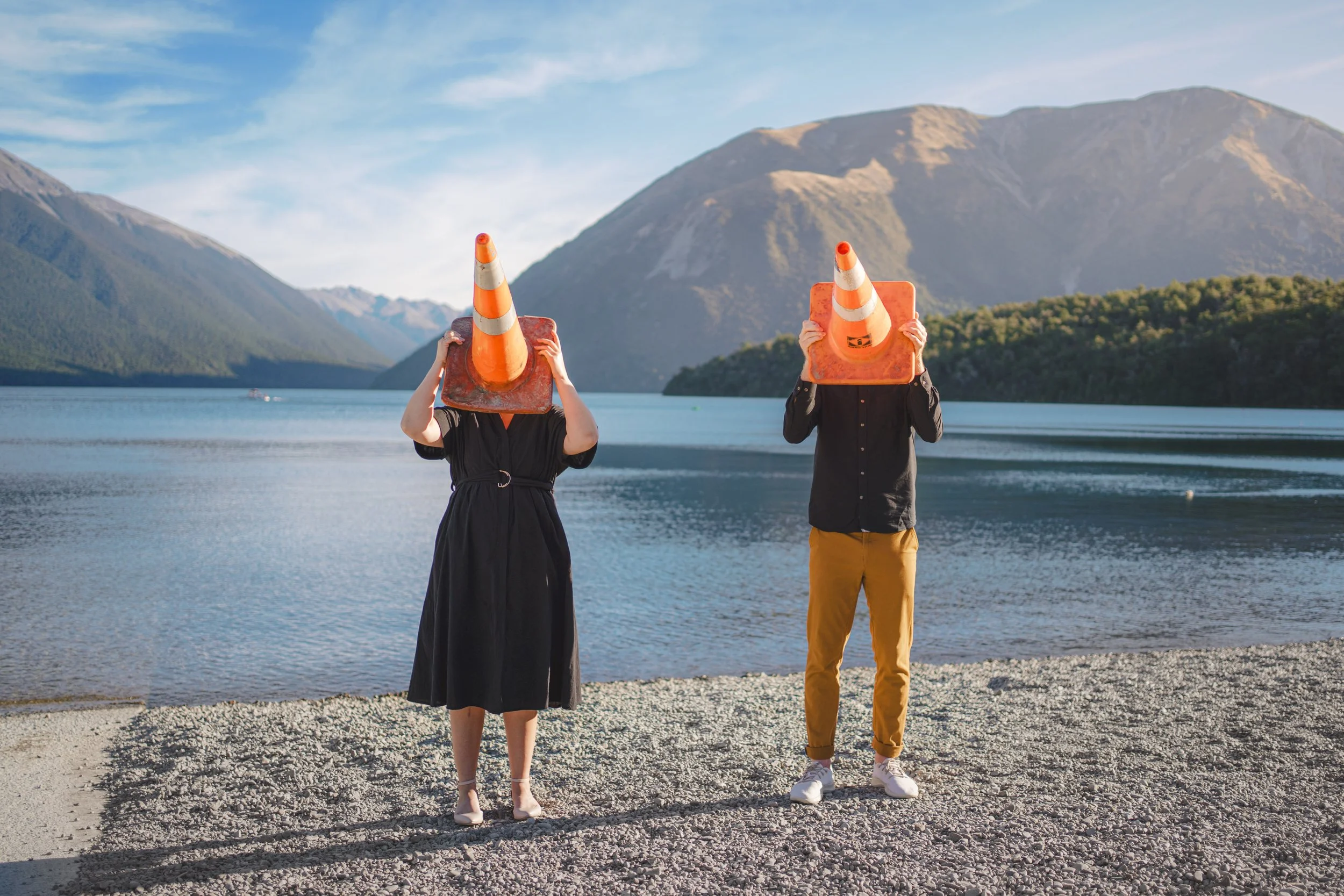 Two people standing on a lakeshore holding traffic cones over their heads like masks, with mountains and a lake in the background.