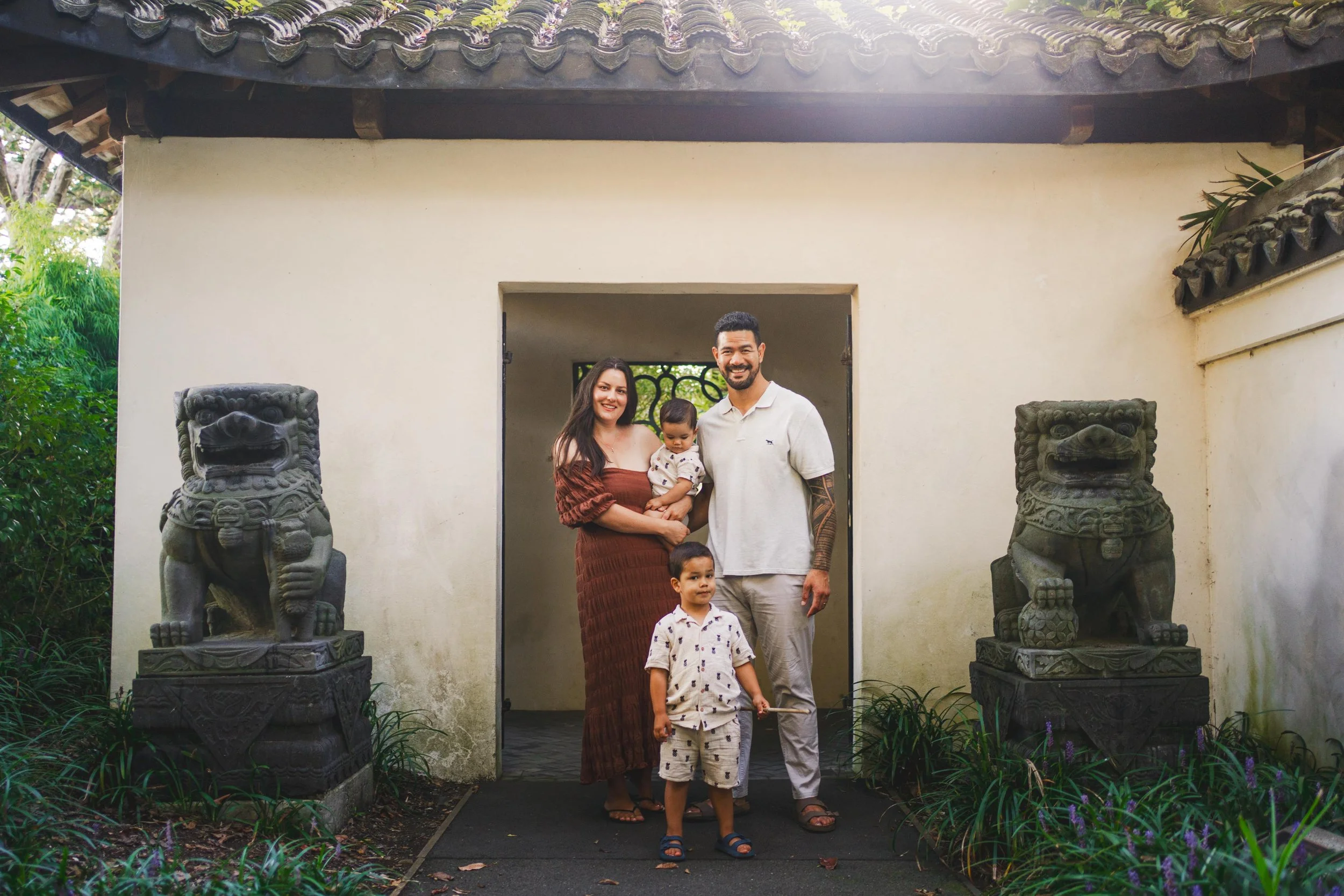 A happy family of four standing in front of a traditional Asian-style building entrance with two stone lion statues on either side. The family includes a woman in a brown dress, a man with a beard in a white polo shirt, a young boy in a beige shirt, 