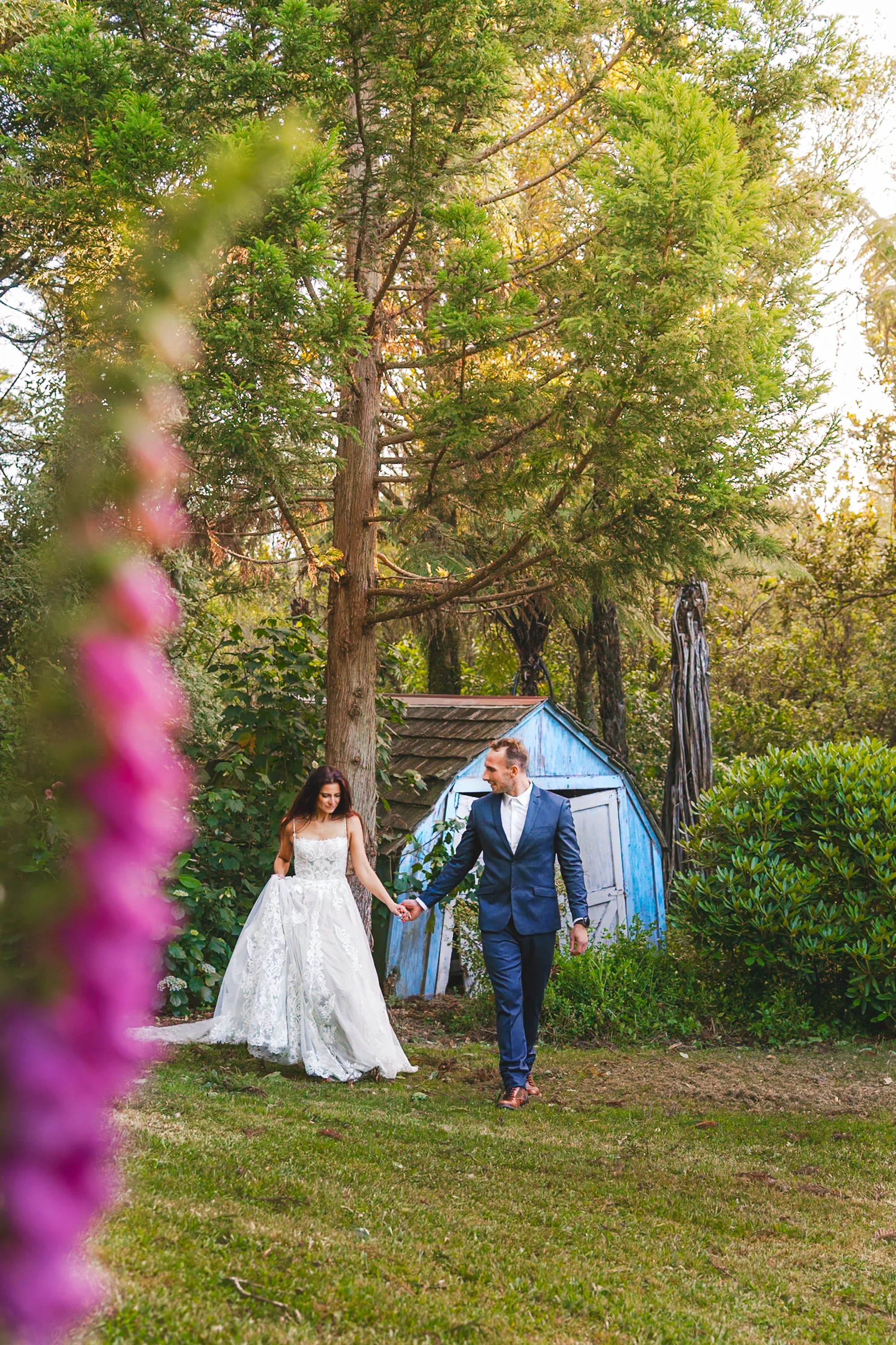 A bride and groom holding hands and walking outdoors in a garden setting near a small blue shed, surrounded by green trees and bushes, with a blurred pink flower in the foreground.