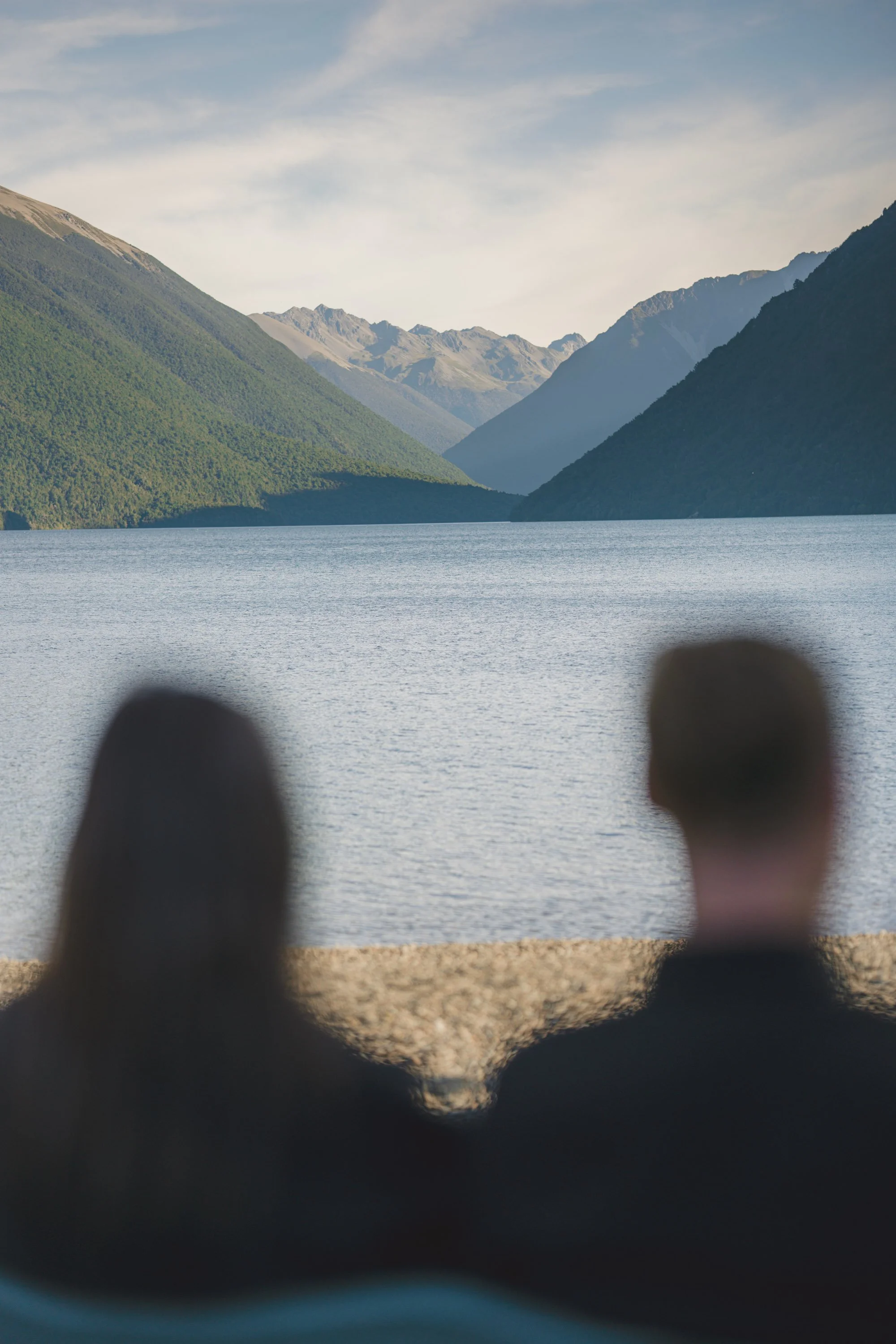 Two people sitting on a rocky shore, looking out at a lake surrounded by green mountains and distant peaks under a partly cloudy sky.