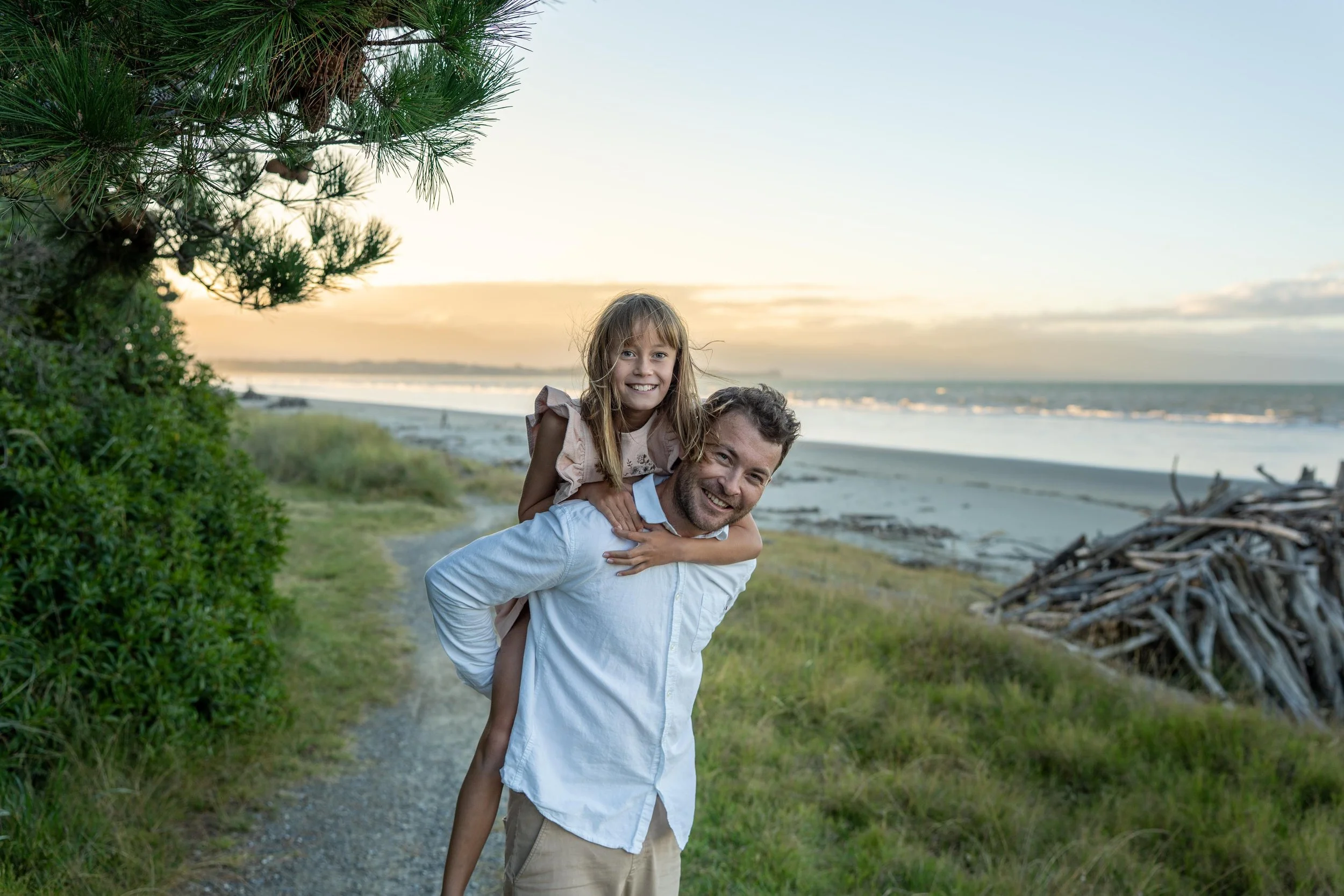A man giving a piggyback ride to a young girl on a beach trail at sunset, with the ocean in the background and greenery on the sides.