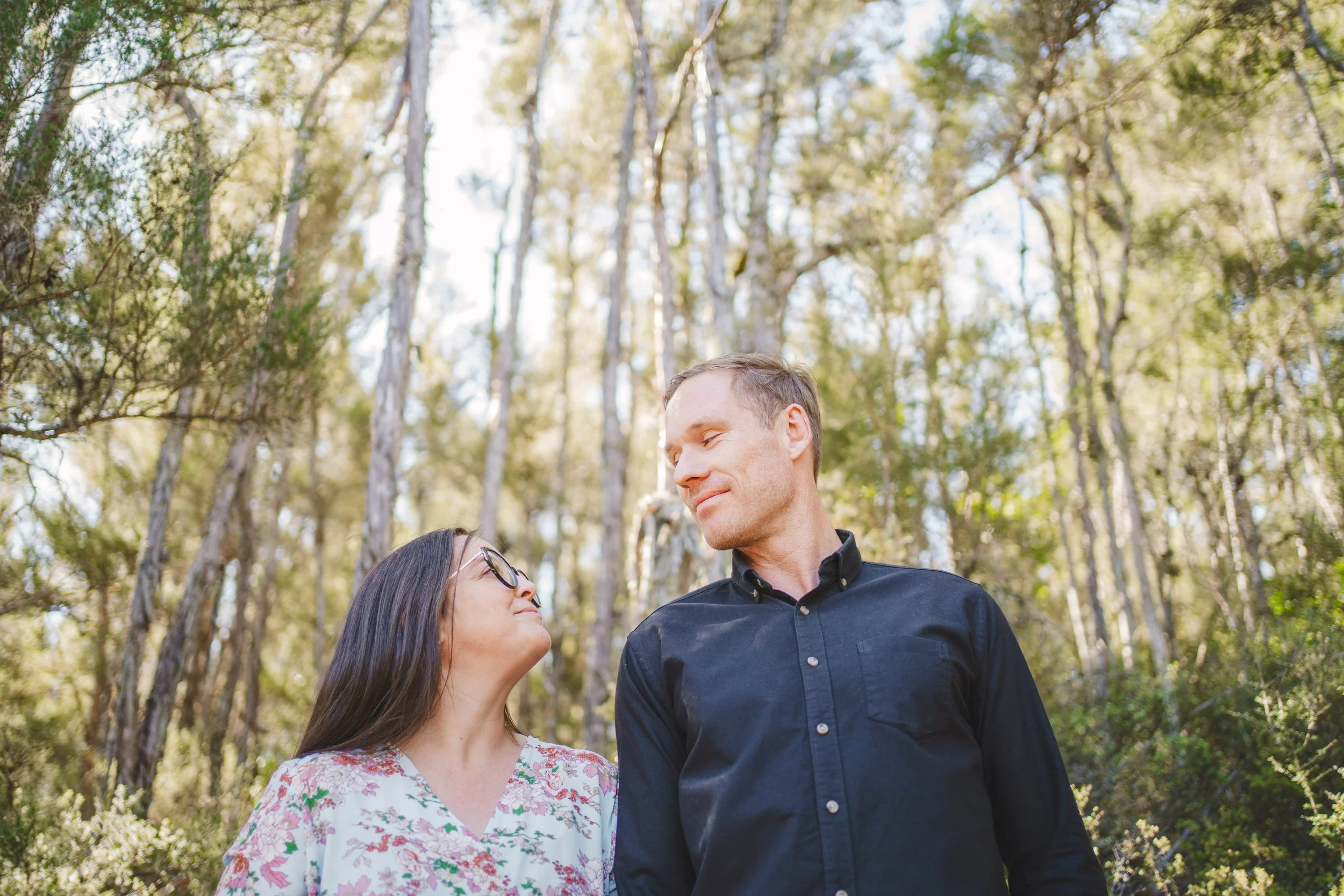 A woman and a man standing close together in a forest, looking at each other lovingly.