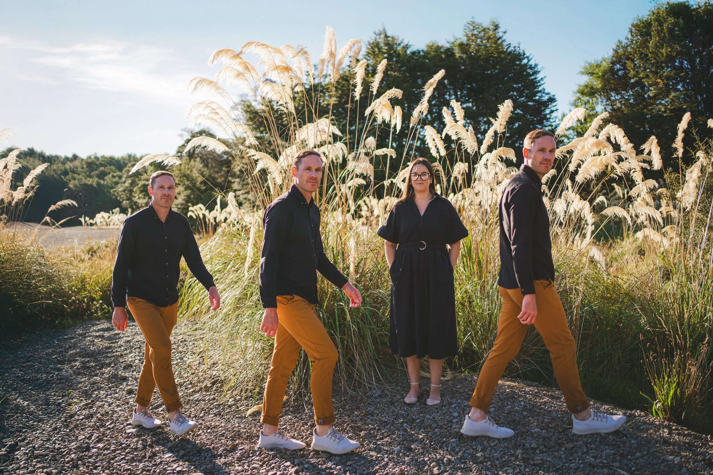 Four people walking on a gravel path in a field of tall grass and plants, with trees in the background and a clear blue sky.