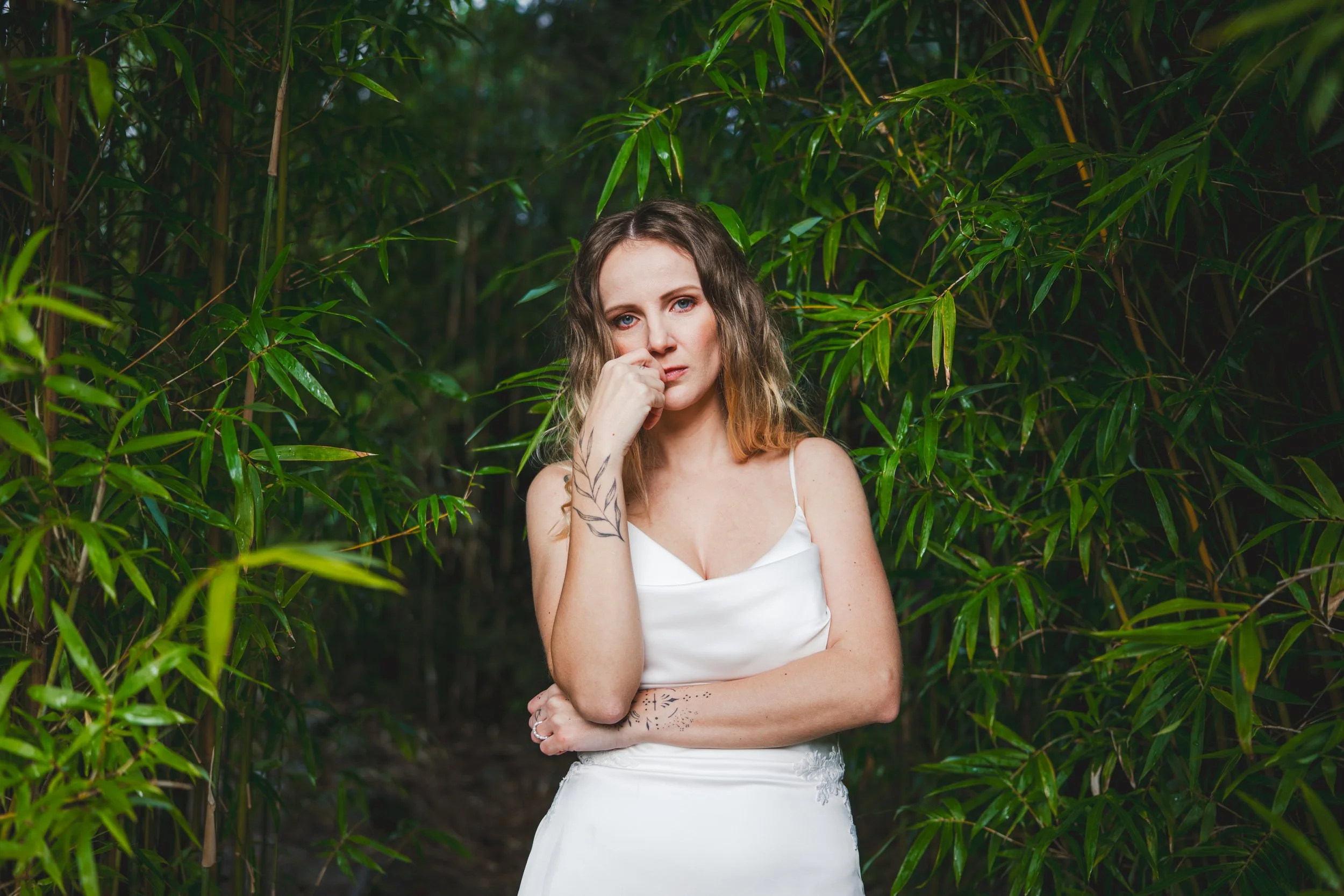 A woman with wavy hair and tattoos on her arm and side, wearing a white dress, stands among green bamboo plants, looking at the camera with a serious expression.