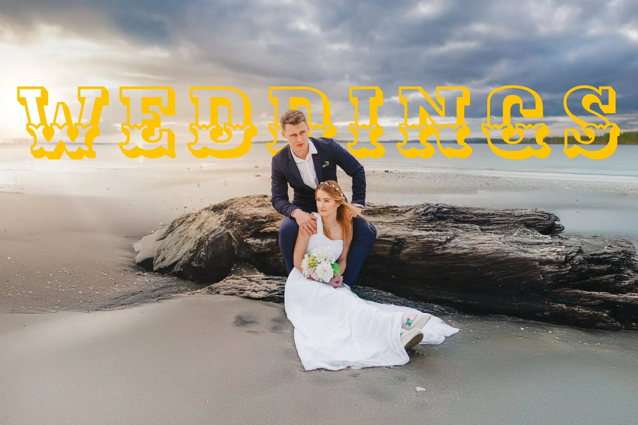 A bride in a white wedding dress sitting on the sand with a bouquet, and a groom in a dark suit kneeling beside her on a large driftwood log at a beach overcast with clouds, with the word 'WEDDINGS' in large yellow letters with a white and yellow outline over the sky.