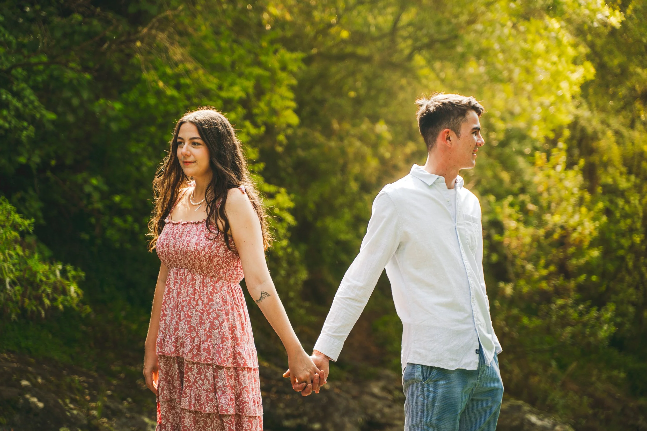 A young woman in a pink floral dress and a young man in a white shirt hold hands while standing outdoors in a lush green area during daytime.