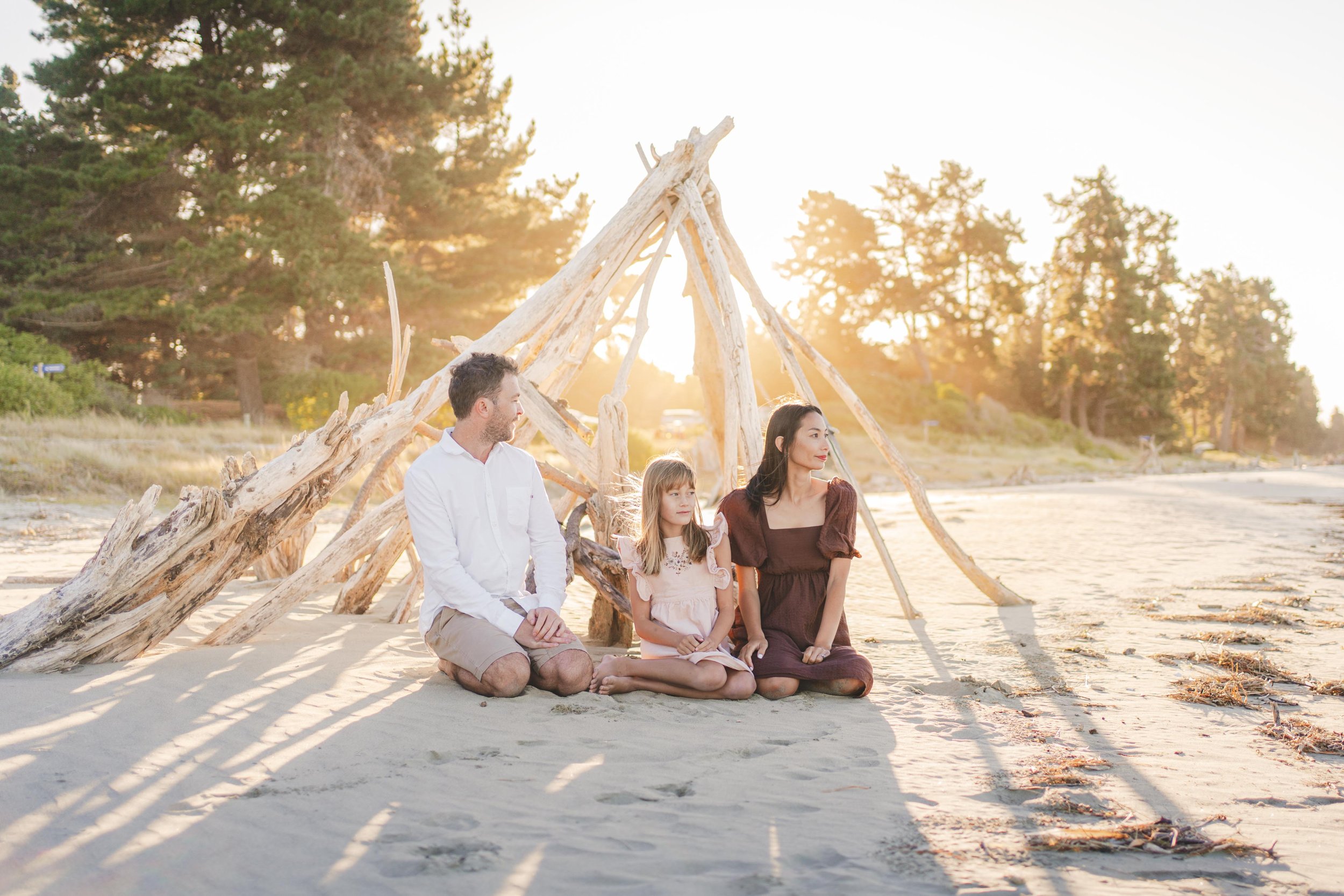 Family of three sitting on a sandy beach near a driftwood teepee at sunset, with trees in the background.
