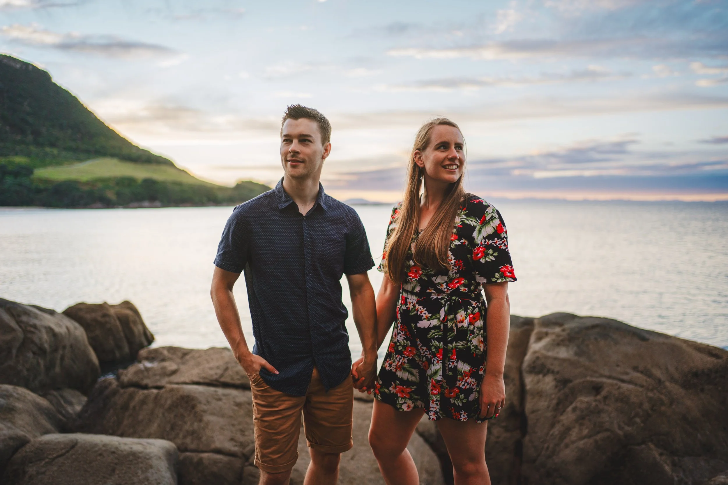 A young couple holding hands by the water at sunset, with a rocky shoreline, a mountain, and a partly cloudy sky in the background.