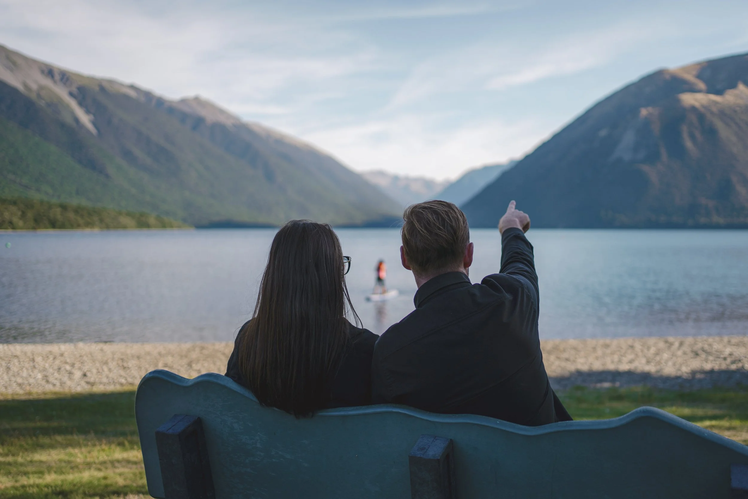 A man and woman sitting on a bench by a lake, with mountains in the background, as one of them is pointing towards a person paddleboarding on the water.