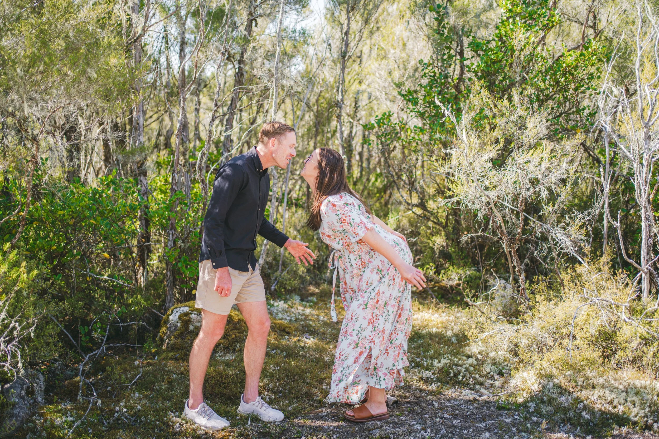 Couple on a forest trail, mischievously confronting each other, with the woman pregnant and wearing a floral dress, and the man in a black shirt and beige shorts.